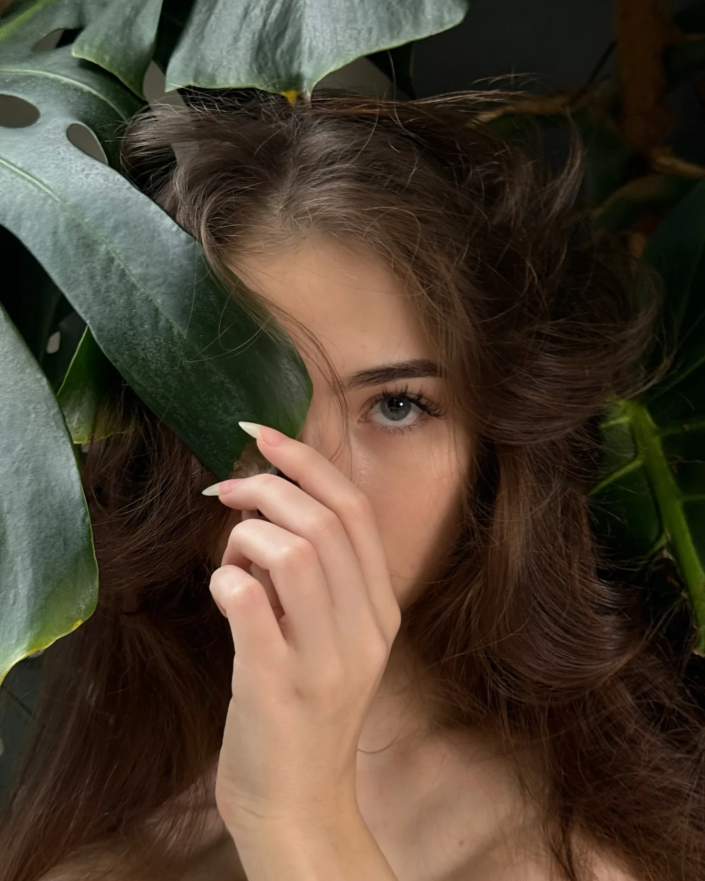 A young woman with brown hair and blue eyes peeks from behind a large green leaf, her hand gently touching her face in a natural, serene portrait.