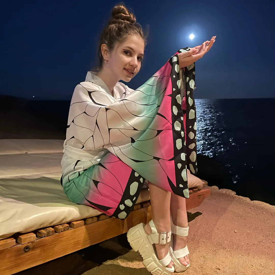 Dana poses on a wooden platform by the sea at night, wearing a colorful butterfly wing patterned dress and white platform sandals, with the moon shining in the background.