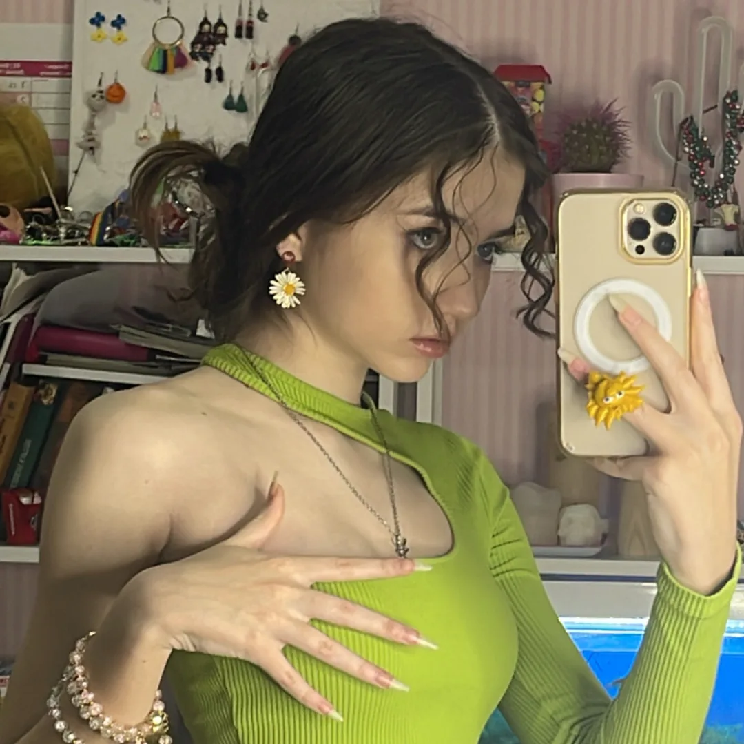 Young woman with dark hair in a ponytail wearing a green top and daisy earrings takes a mirror selfie with a gold iPhone featuring a yellow flower grip.