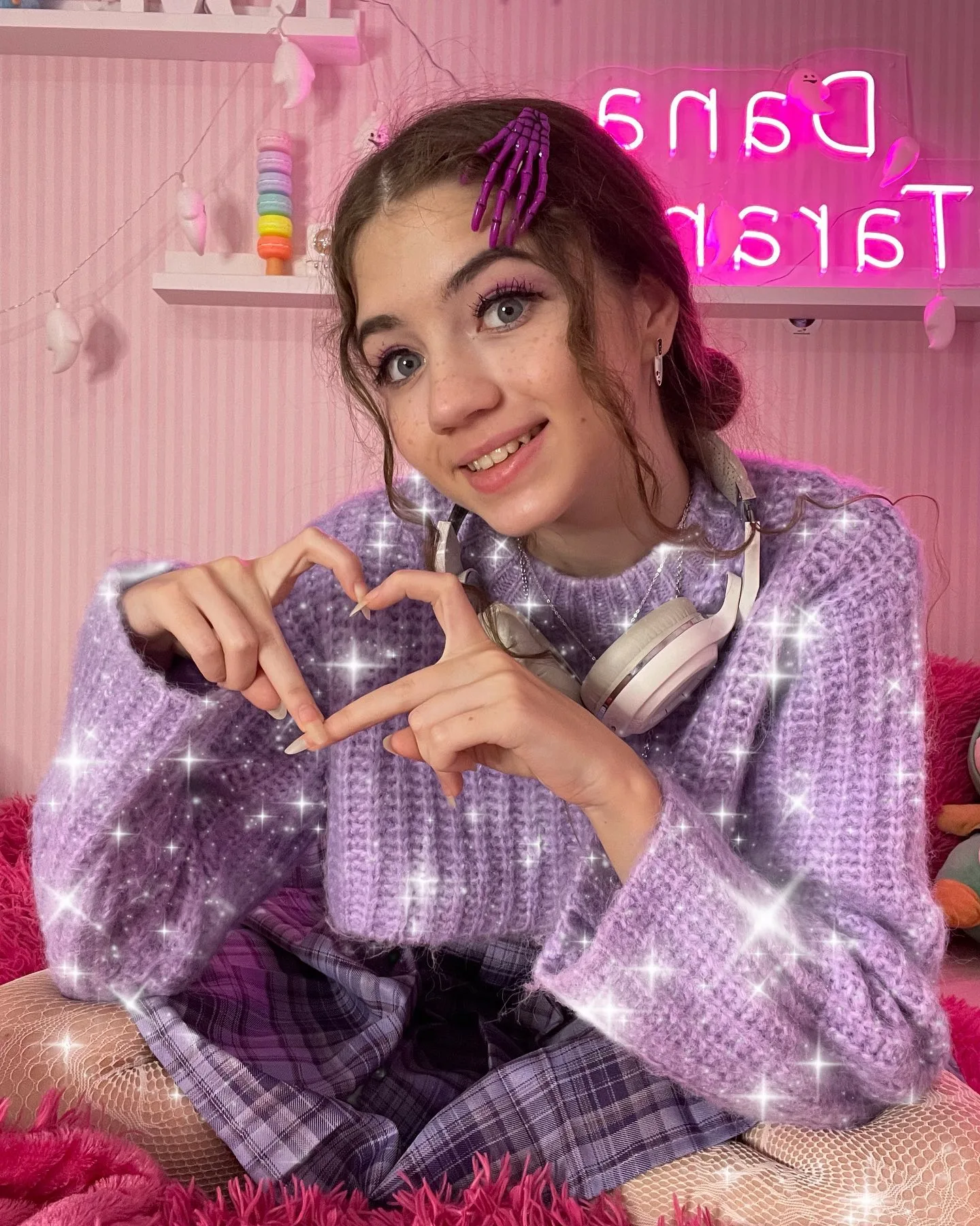 Dana poses in a purple sweater making a heart shape with her hands, with a pink neon sign and headphones visible in the background.