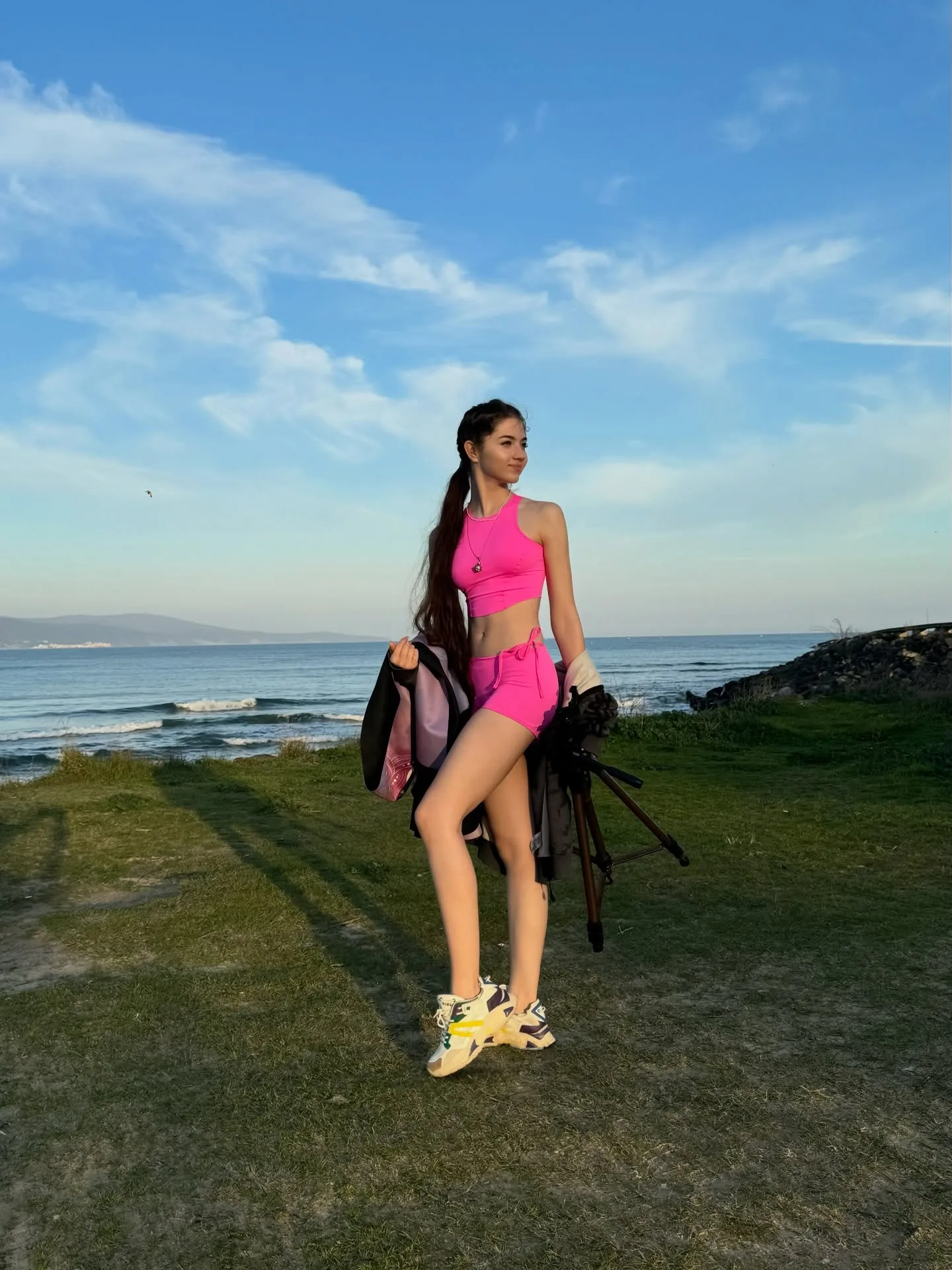 Woman in pink sportswear standing on grassy cliff overlooking the ocean with a blue sky.