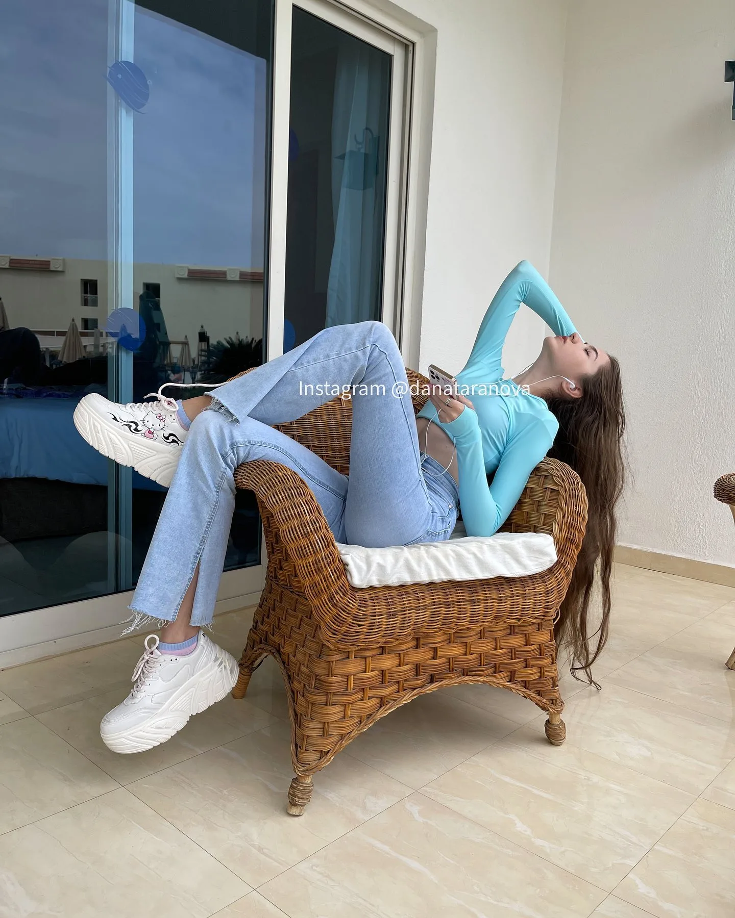 A woman in a light blue long-sleeve top and jeans reclines in a wicker chair with white sneakers, posing near a glass door.