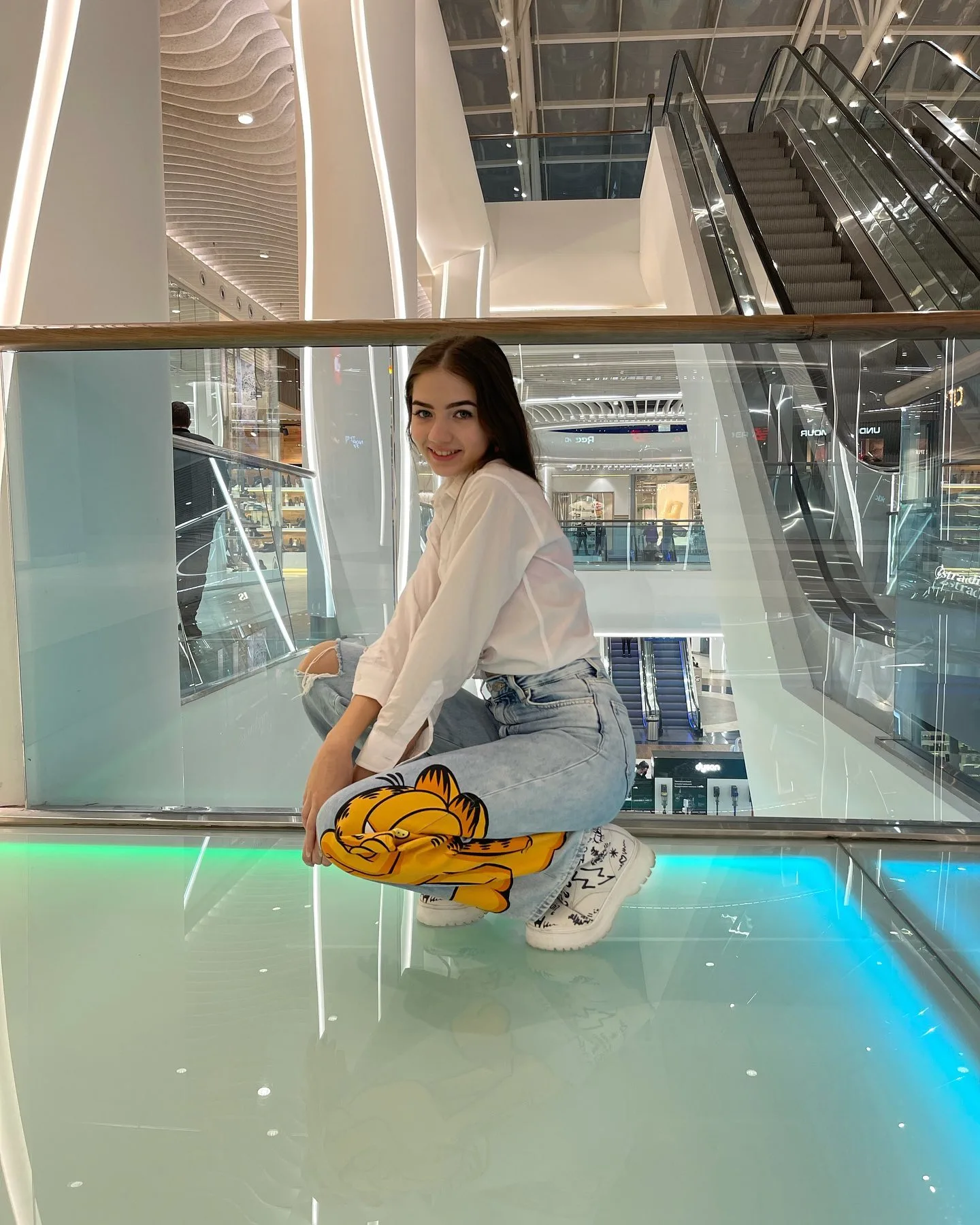 A woman in a white top and yellow graphic jeans crouching on a glass floor in a modern shopping mall with escalators in the background.