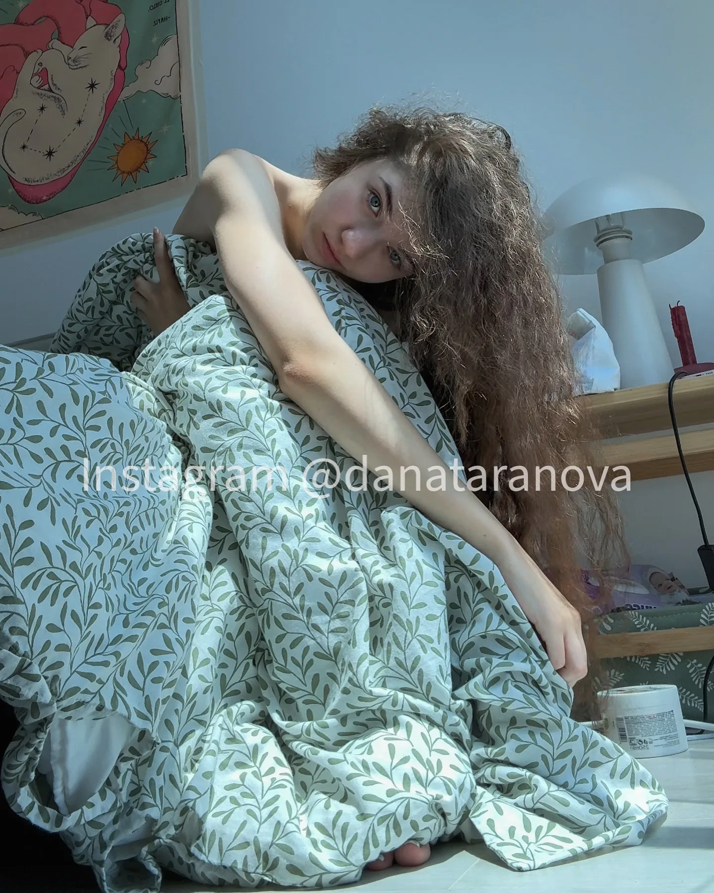 Dana resting in bed with curly hair, wrapped in a leaf-patterned duvet, with a painting and lamp in the background.