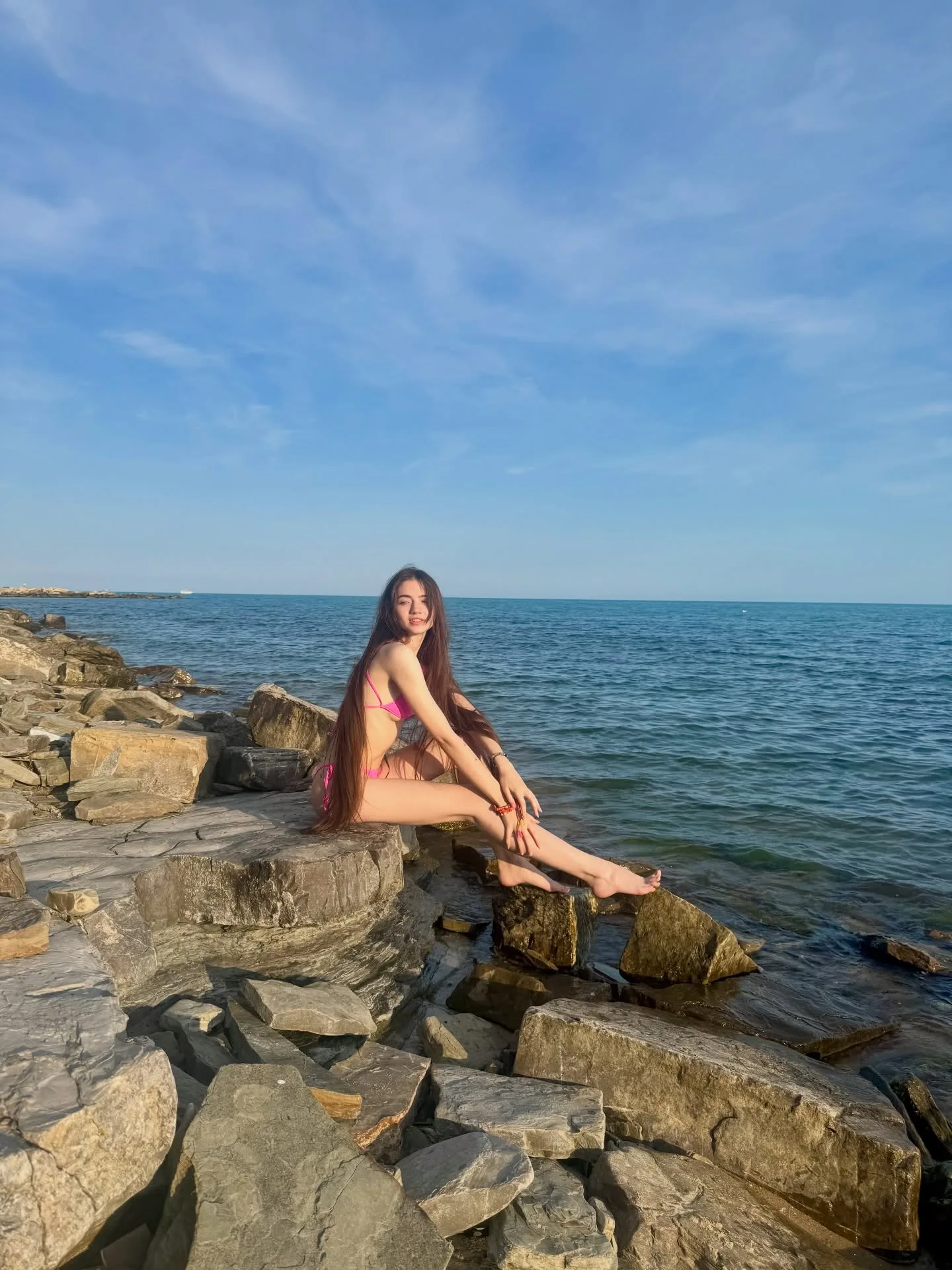 A woman in a pink bikini sits on rocky shoreline rocks with the ocean and blue sky in the background.