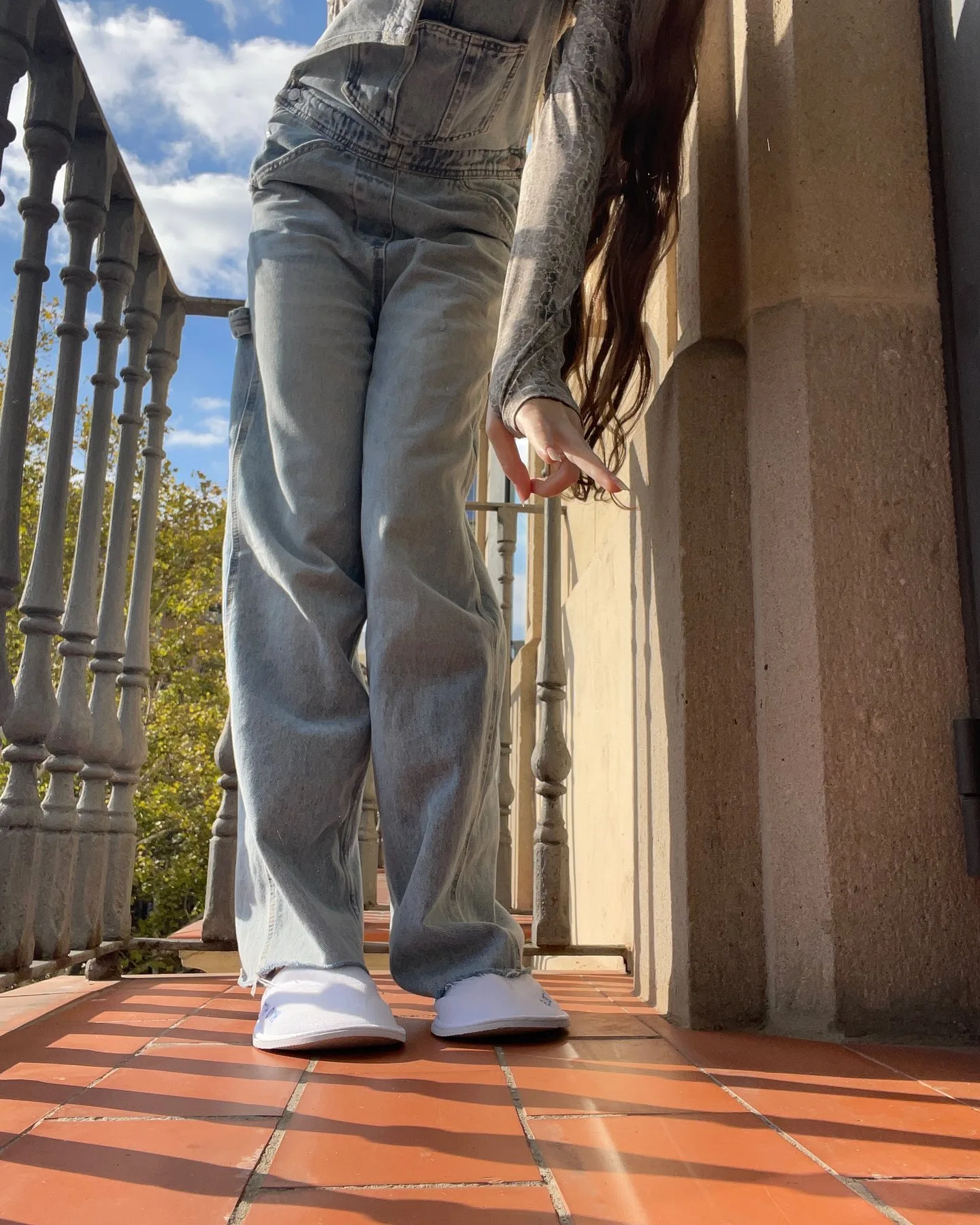 A person wearing light blue jeans and white sneakers stands on a tiled balcony with a metal railing and blue sky.