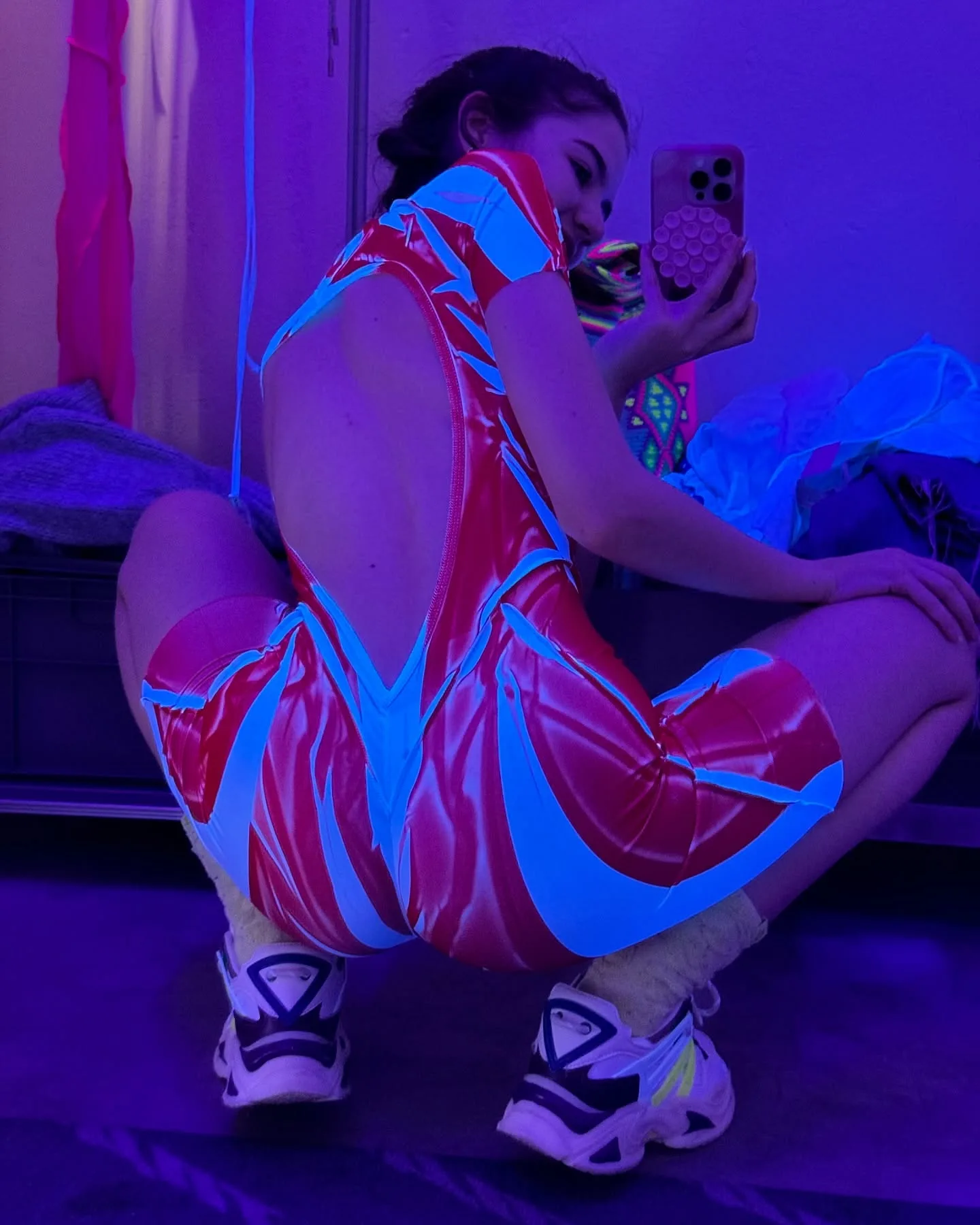 Woman in a red and blue patterned bodysuit taking a mirror selfie in purple lighting.