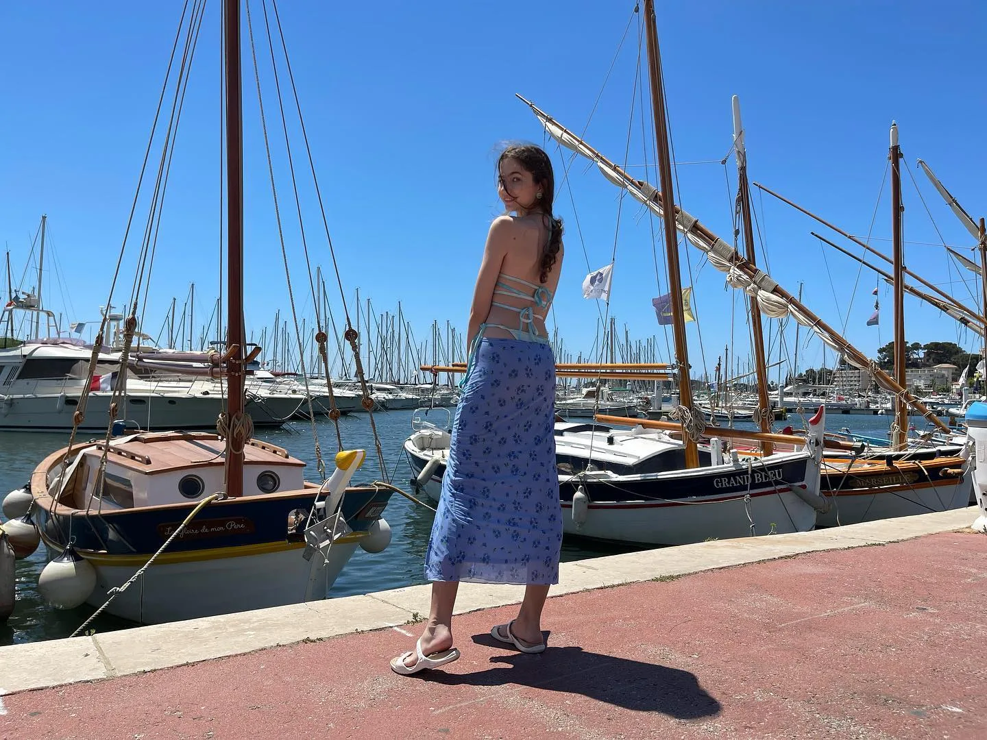 Woman in blue floral skirt standing on a red marina walkway with sailboats in the background.