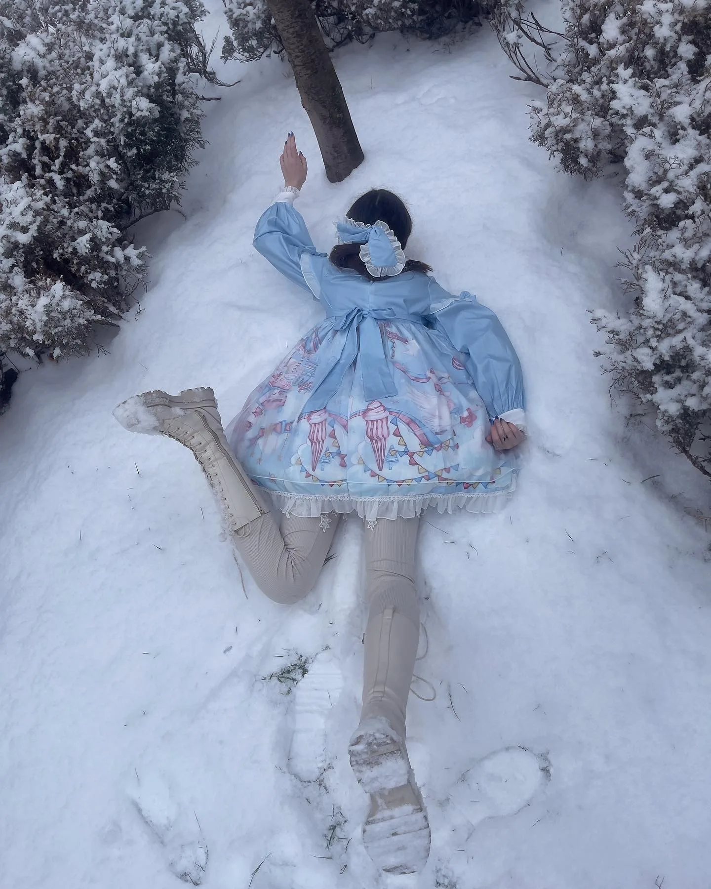 A person in a blue Lolita dress lies on their back in the snow, making a snow angel with arms and legs spread wide.