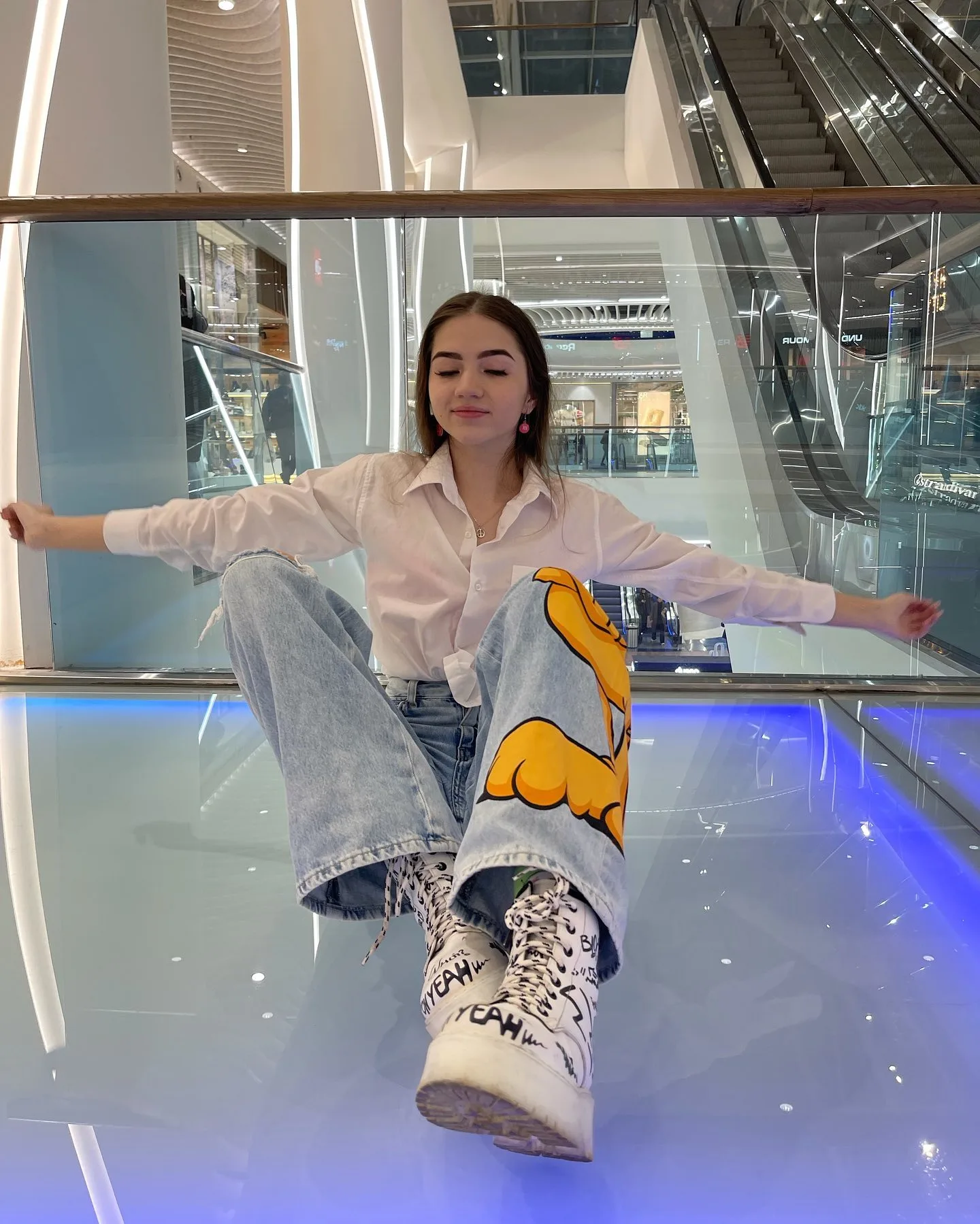 A woman sits on a glass floor in a modern mall wearing a white shirt, light blue jeans with a yellow graphic, and white patterned boots.
