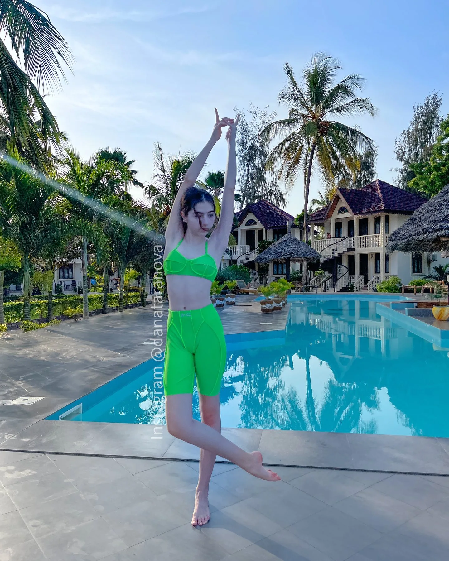 Woman in green bikini and shorts posing by a pool with palm trees and resort buildings in the background.