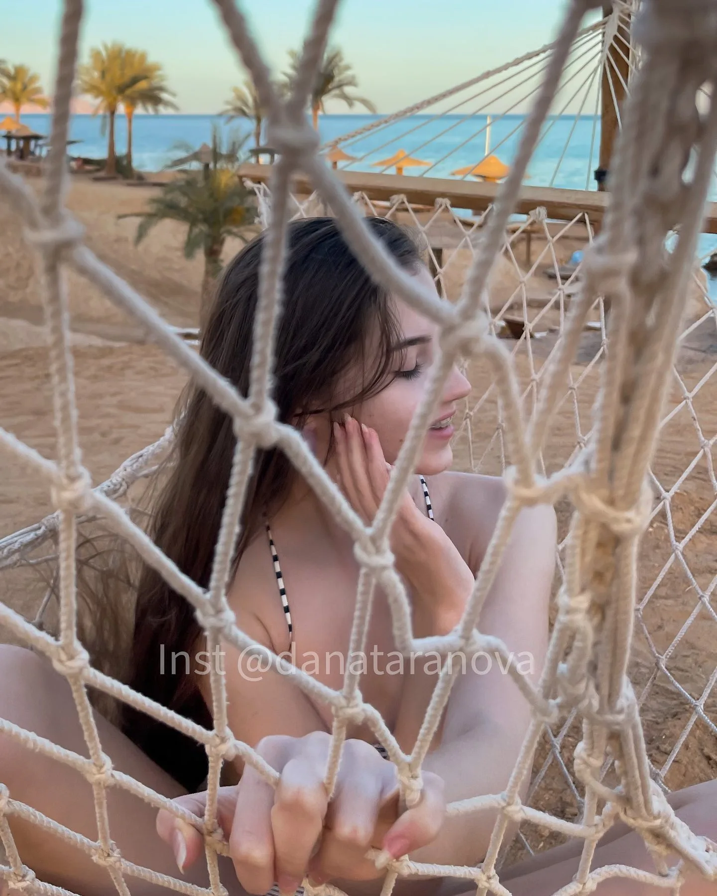 A woman with long dark hair sits in a rope net hammock on a sandy beach, with palm trees and the ocean visible in the background.