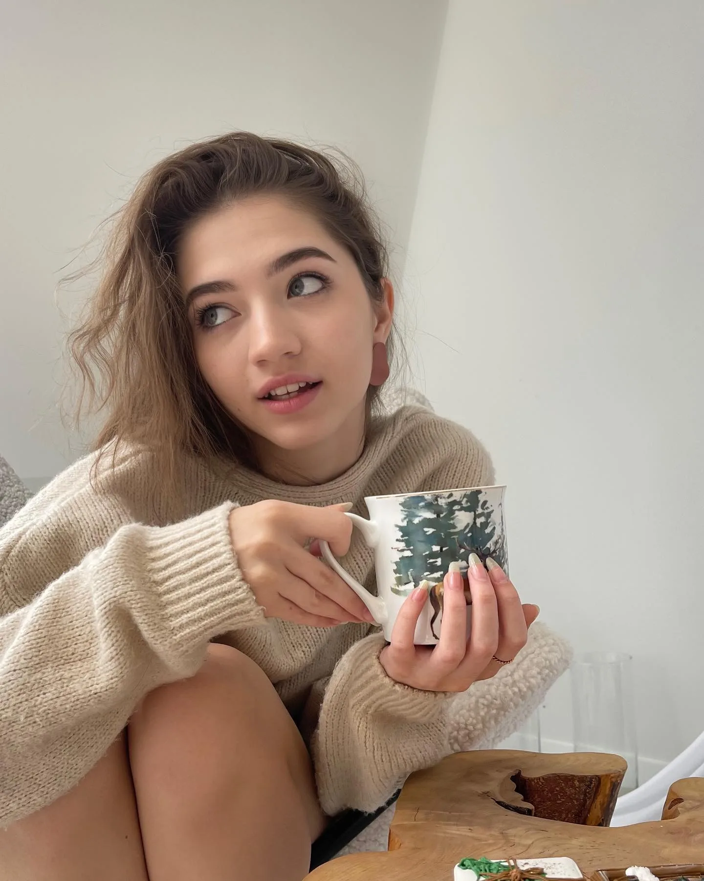 Young woman in a beige sweater holding a mug with a tree design, looking up with a thoughtful expression.