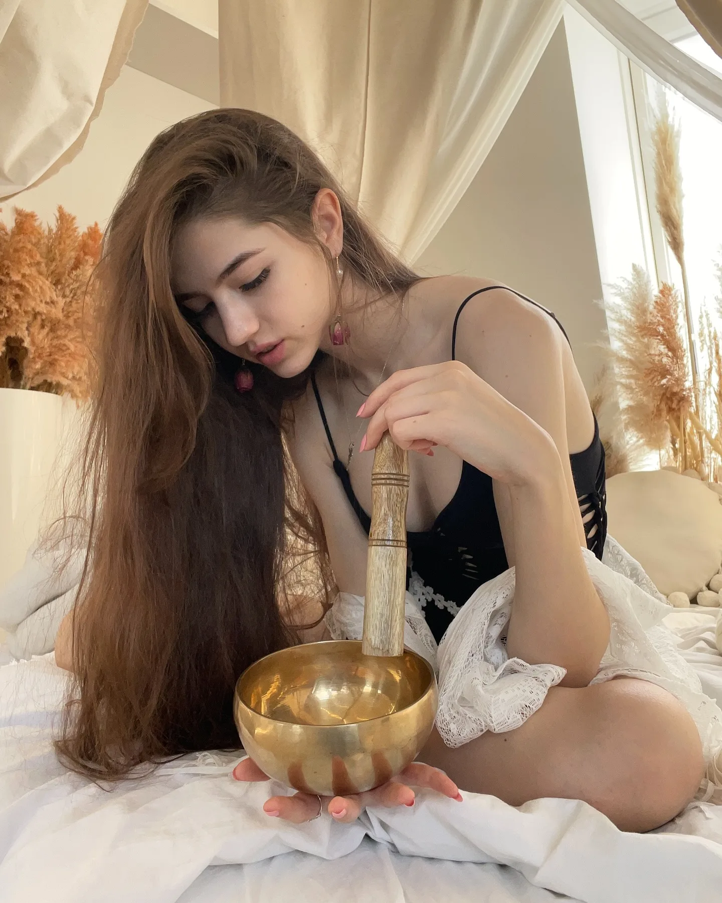 A woman with long brown hair sits on a bed striking a golden singing bowl with a wooden mallet, surrounded by soft lighting and dried pampas grass.
