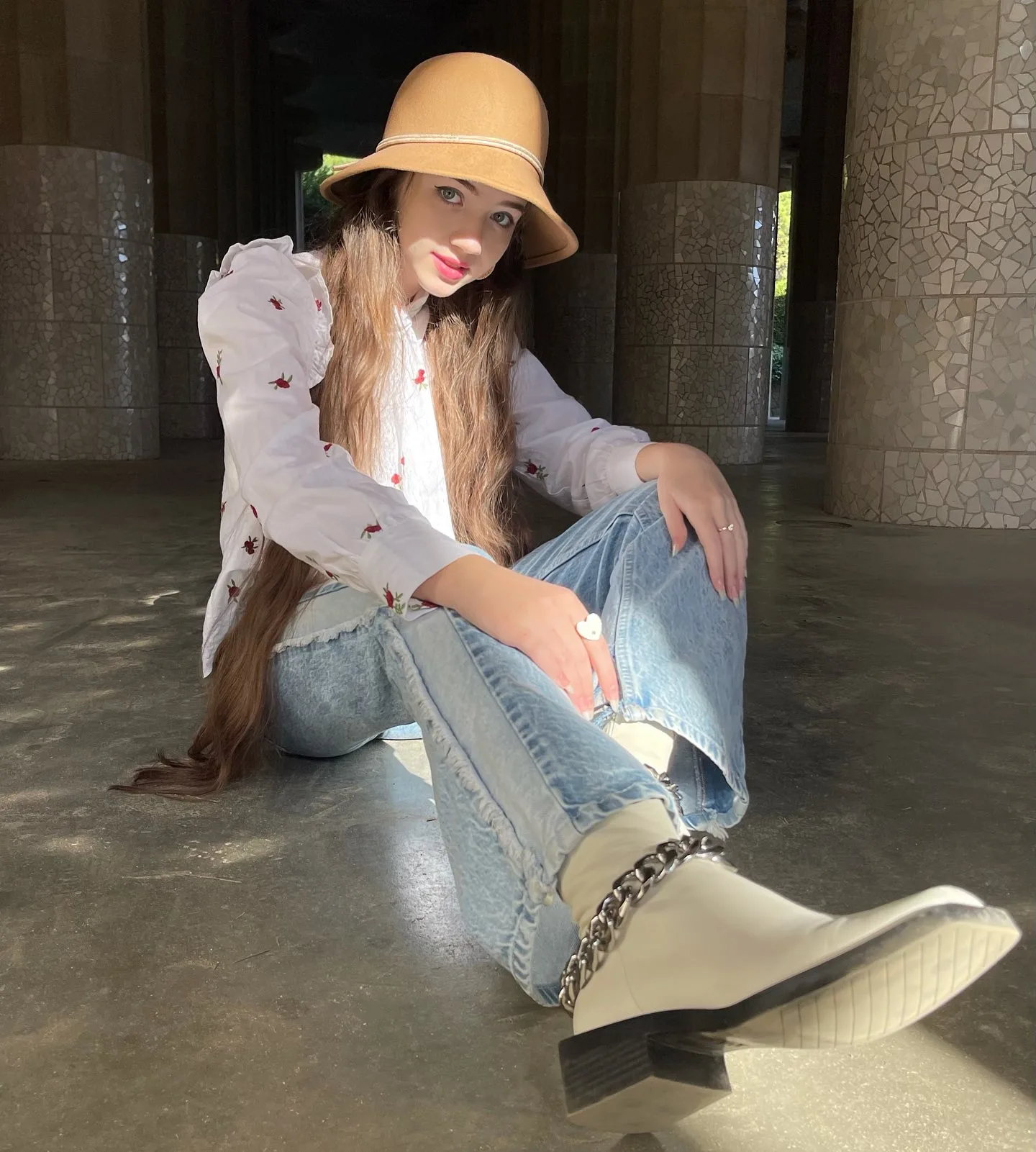 Dana wearing a beige bucket hat, white blouse, and blue jeans sitting on a concrete floor.