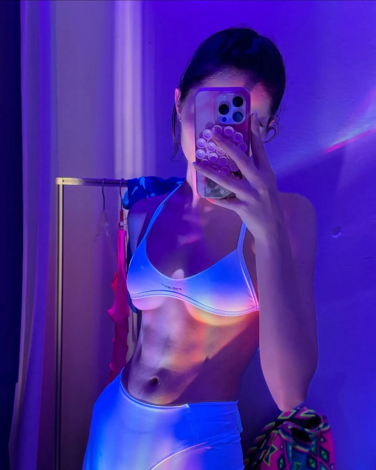 A woman taking a mirror selfie in a colorful bikini top and matching bottoms under purple lighting.