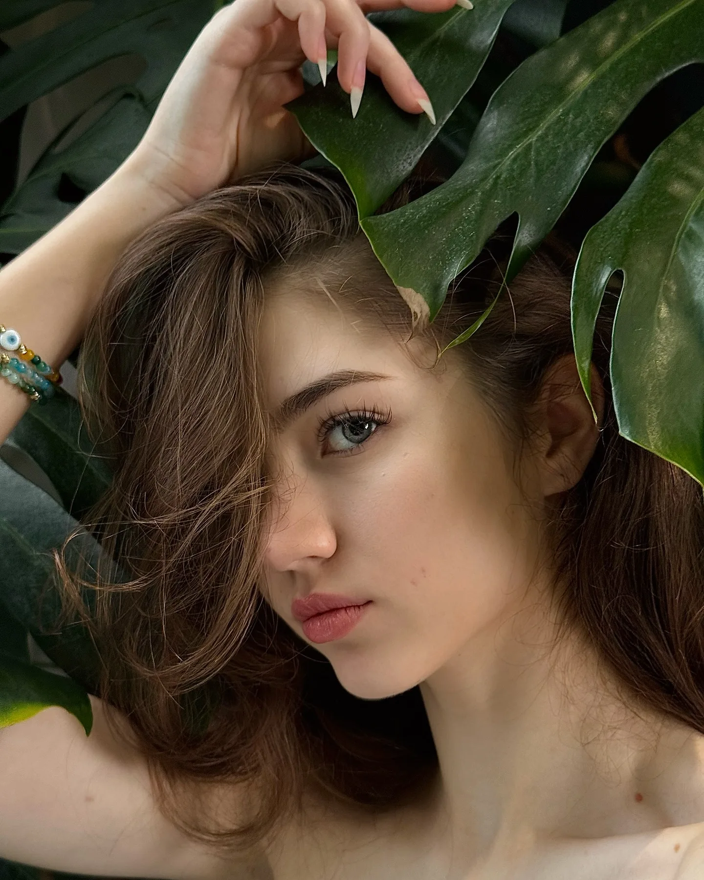 Portrait of a young woman with brown hair and blue eyes, wearing a bracelet, posing among large green monstera leaves.