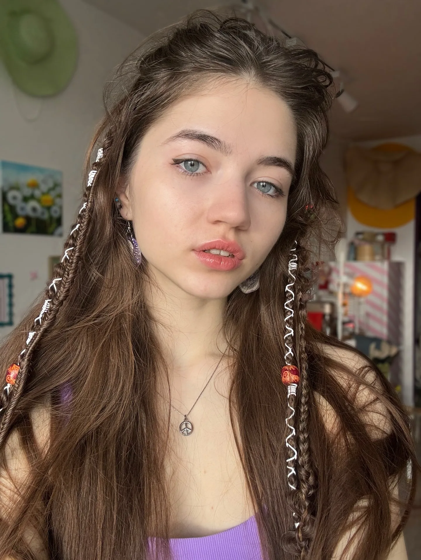 Portrait of a young woman with long brown hair featuring decorative braids and beads, wearing a purple top and necklace.