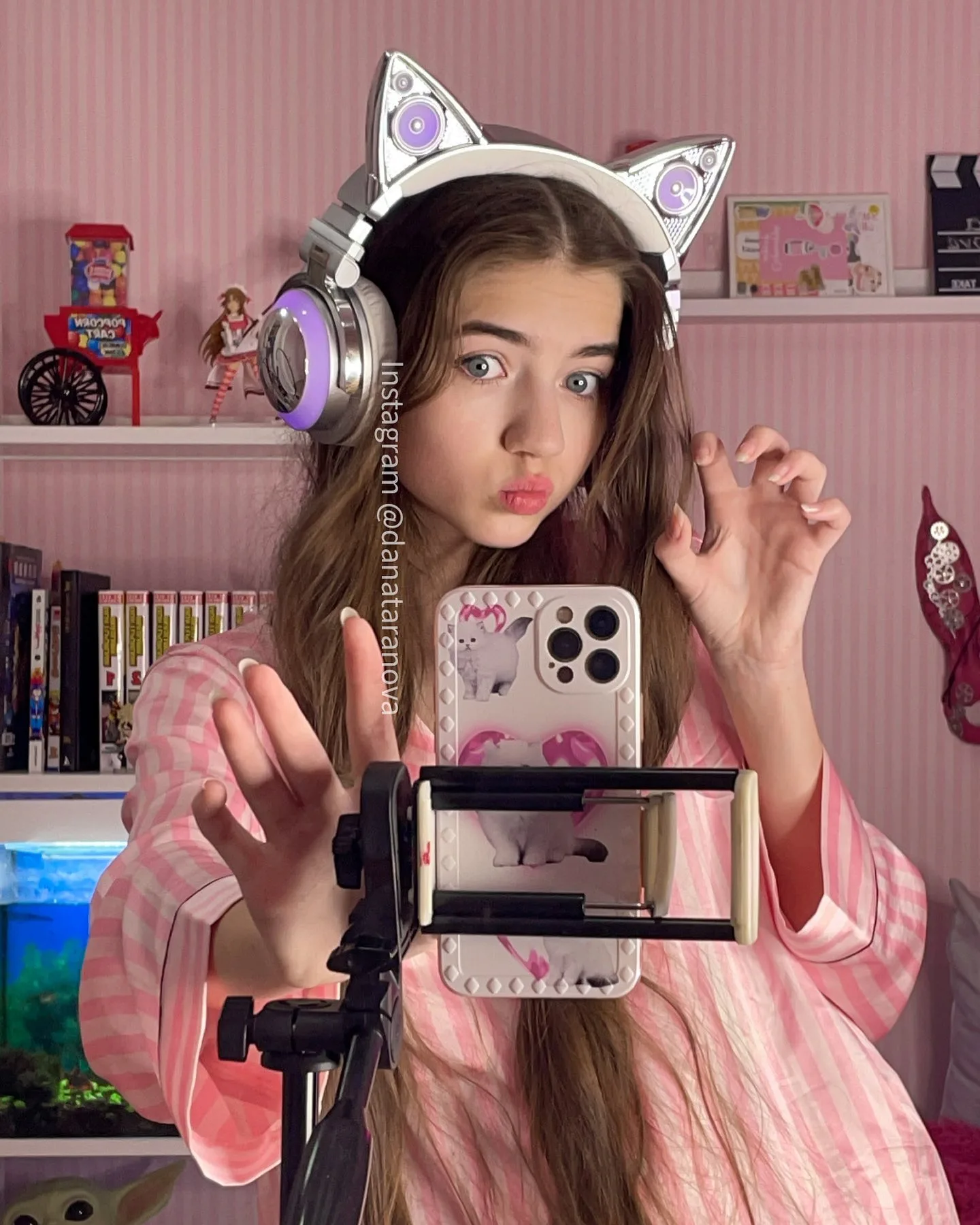 Teenage girl with long brown hair wearing silver cat ear headphones and pink plaid pajamas taking a mirror selfie with a smartphone on a tripod in a pink bedroom.