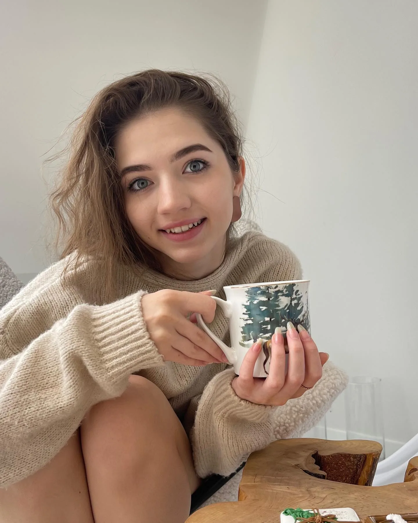 Dana smiling while holding a mug with a forest design, wearing a cozy beige sweater.