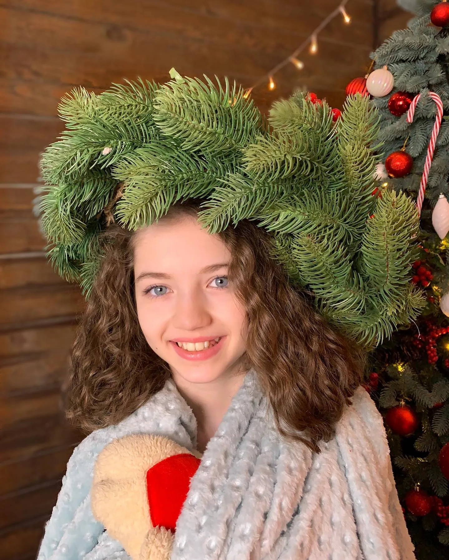 A smiling girl with curly hair wears a large pine branch crown and holds a teddy bear near a decorated Christmas tree.
