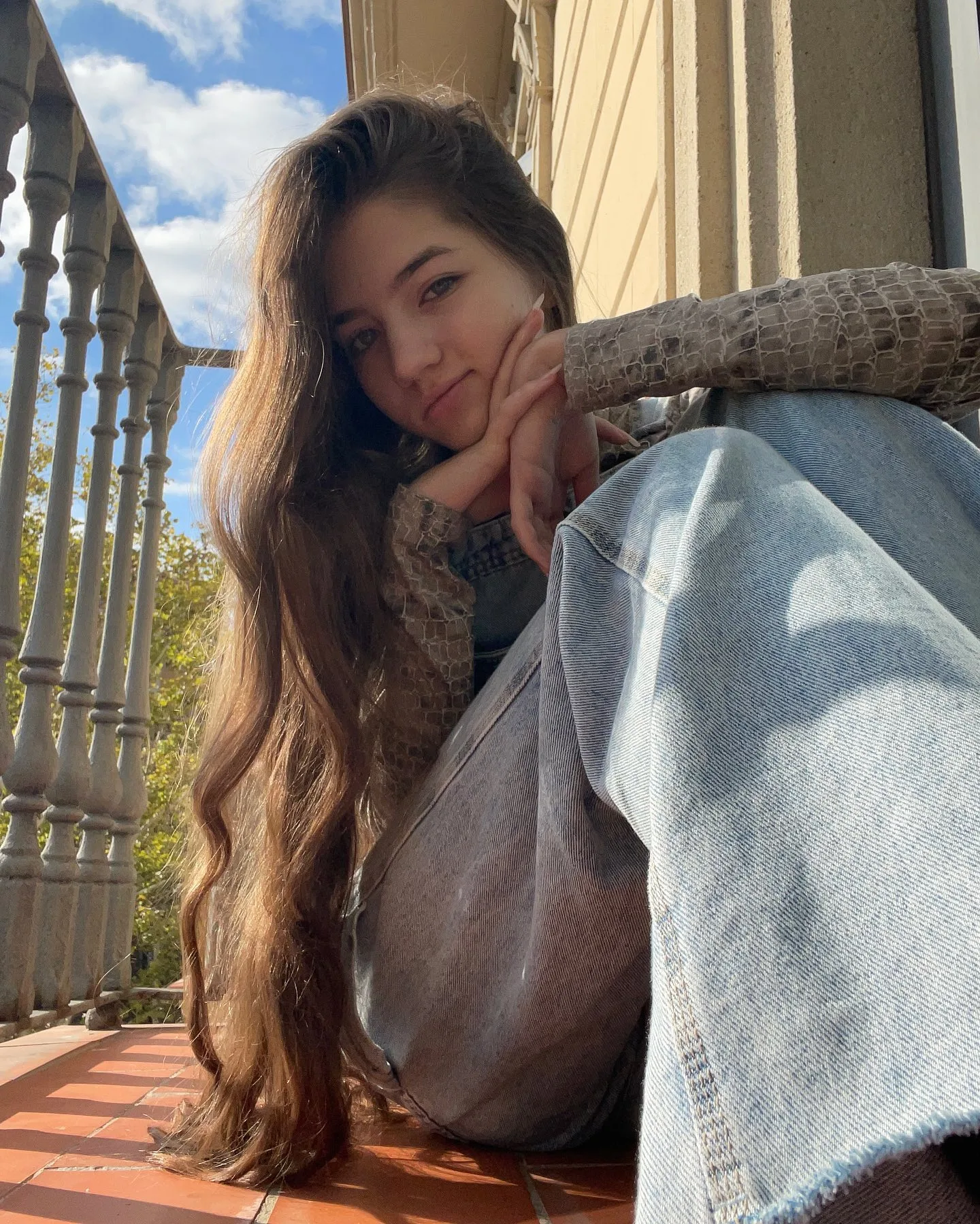 Young woman with long wavy brown hair sitting on a wooden balcony railing, wearing a beige sweater and light blue jeans, resting her chin on her hand against a blue sky.