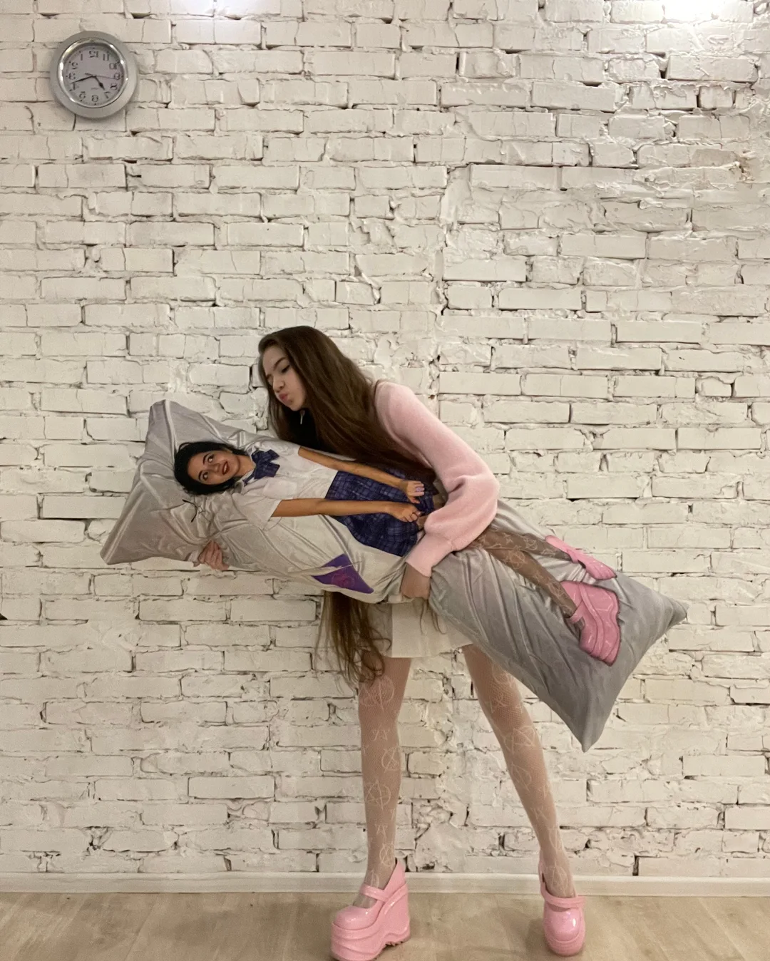 A woman in a pink sweater and fishnet tights hugs a large body pillow featuring a man's face against a white brick wall.