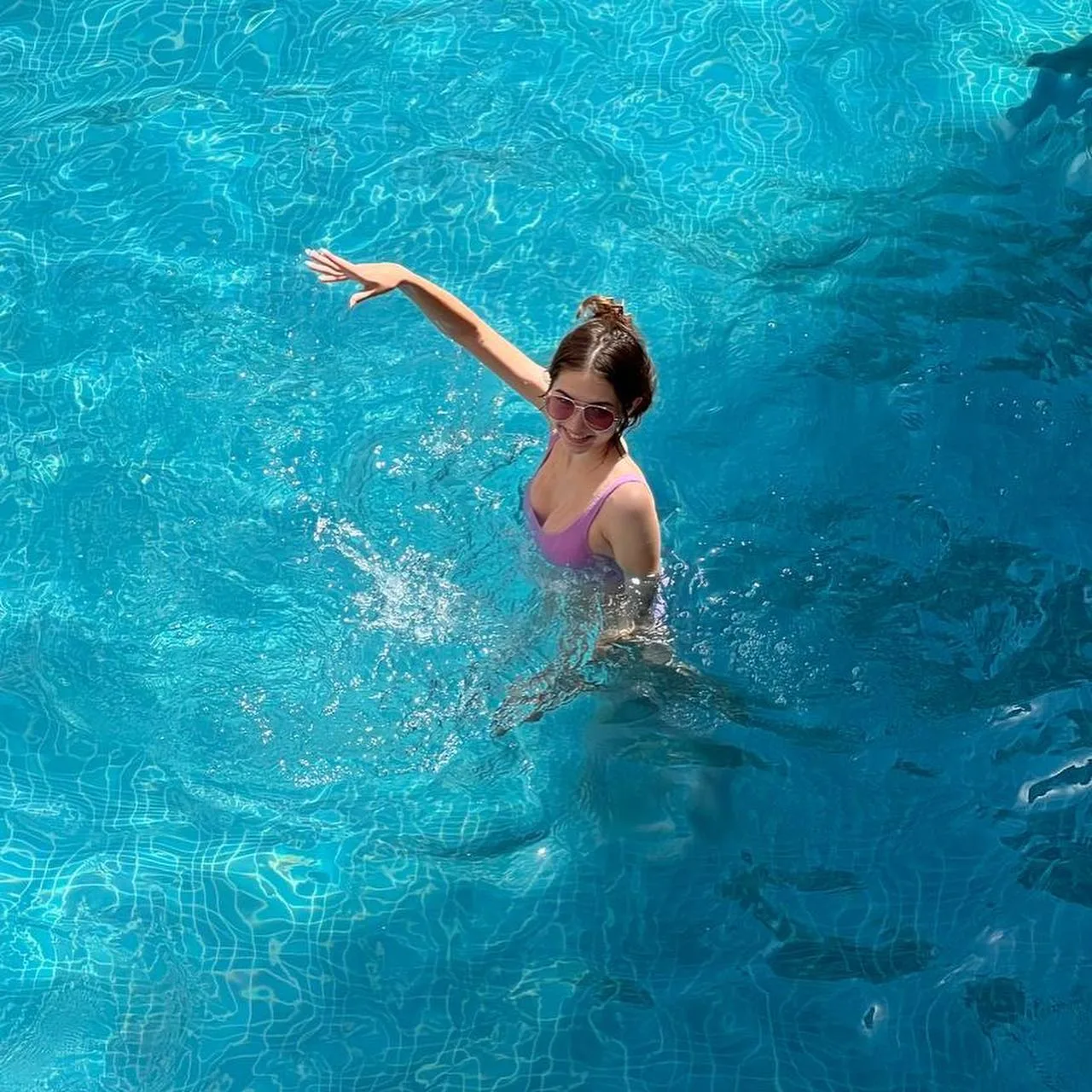 A woman in a pink swimsuit and sunglasses poses in a blue swimming pool with her arm raised.