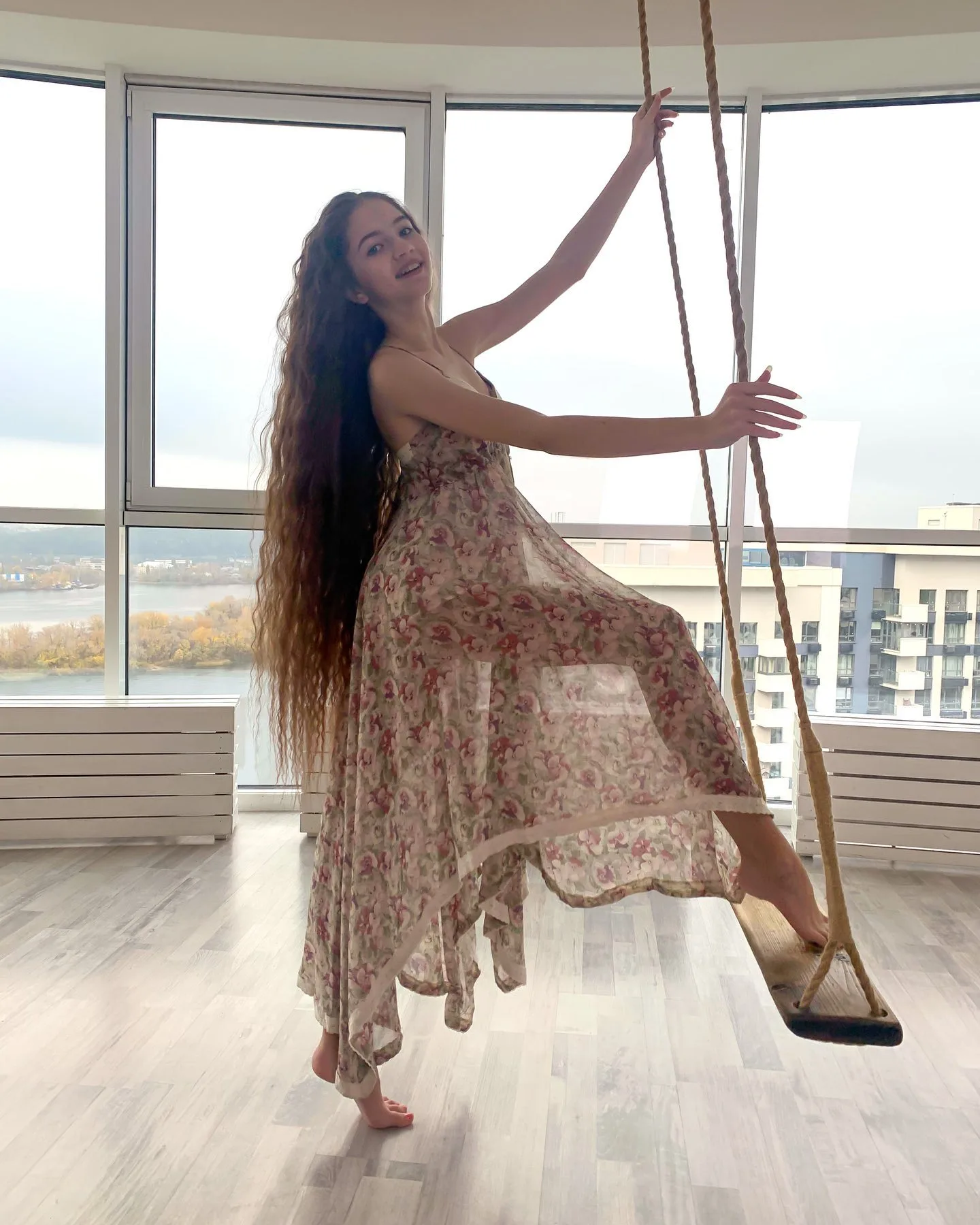 A woman with long hair in a floral dress poses on an aerial silk swing in a bright room with large windows.
