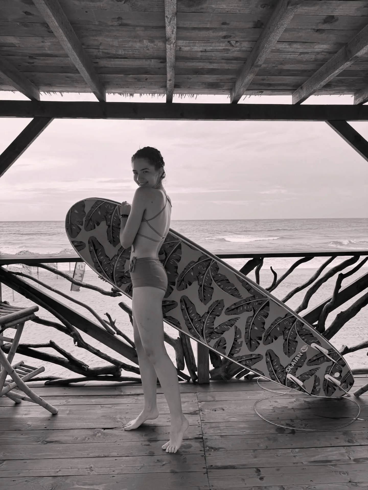A woman in a swimsuit stands on a wooden deck holding a surfboard with a leaf pattern, looking out at the ocean.