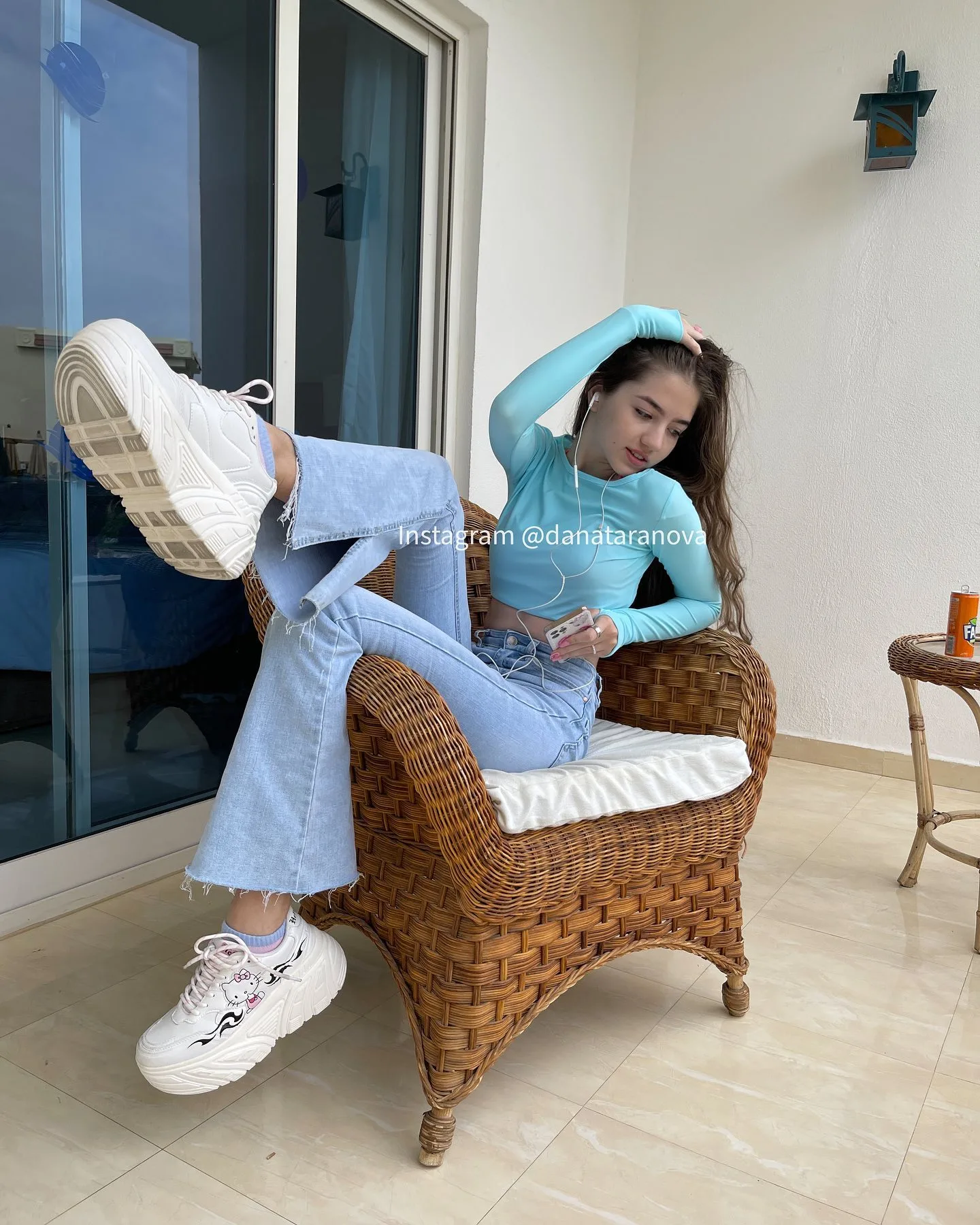 A young woman with long brown hair sits on a wicker chair wearing a light blue long-sleeved top and light blue jeans, posing with one leg raised and white sneakers visible.