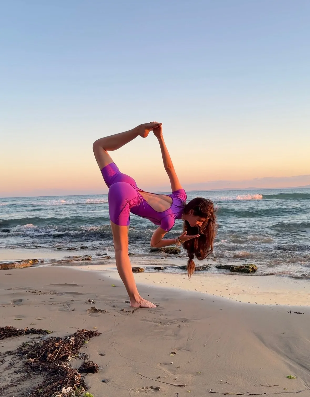 Woman in purple swimsuit performing a standing split yoga pose on a sandy beach at sunset.