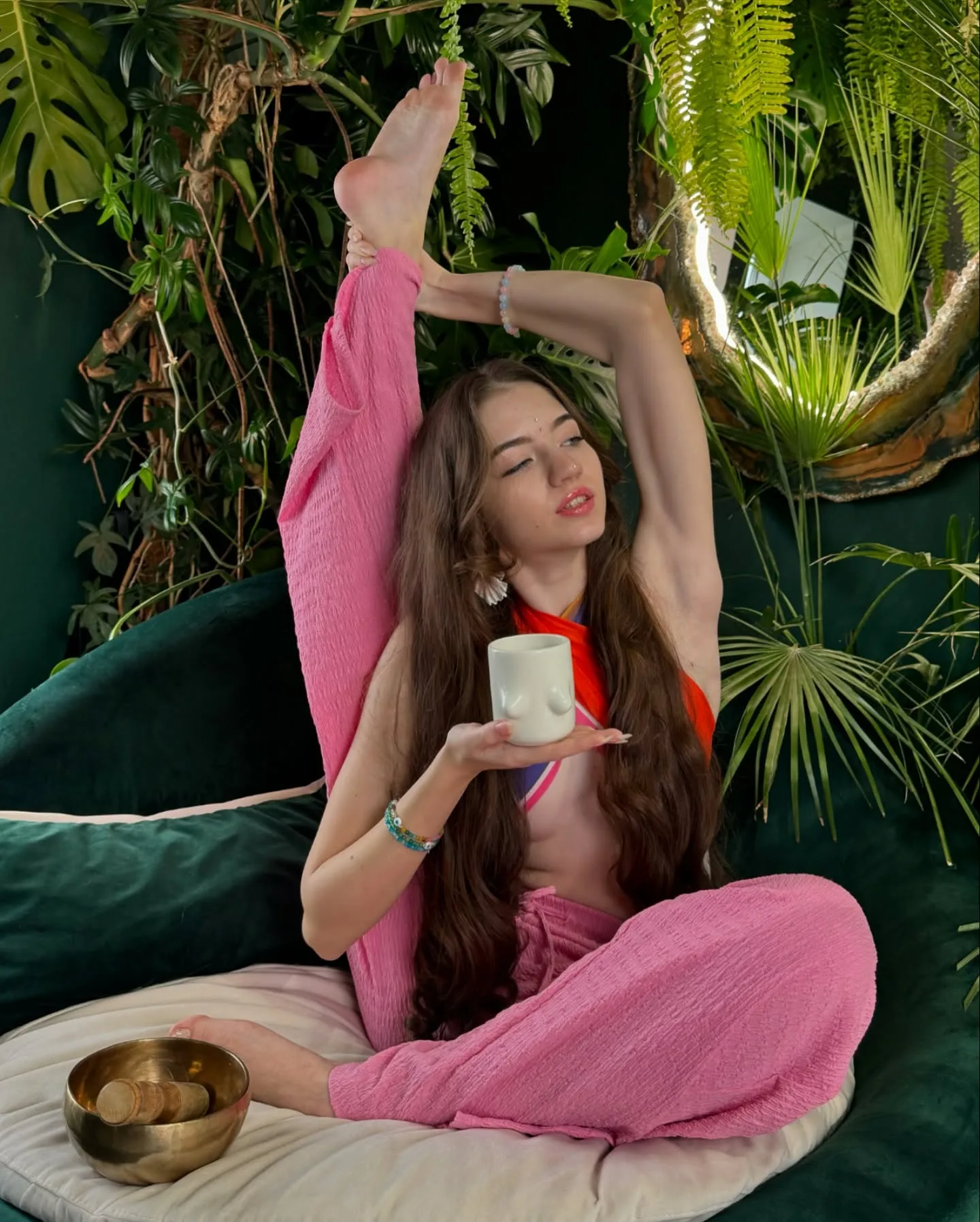 Woman in pink yoga pants performing a seated leg stretch while holding a white mug, surrounded by lush green plants and a golden singing bowl.