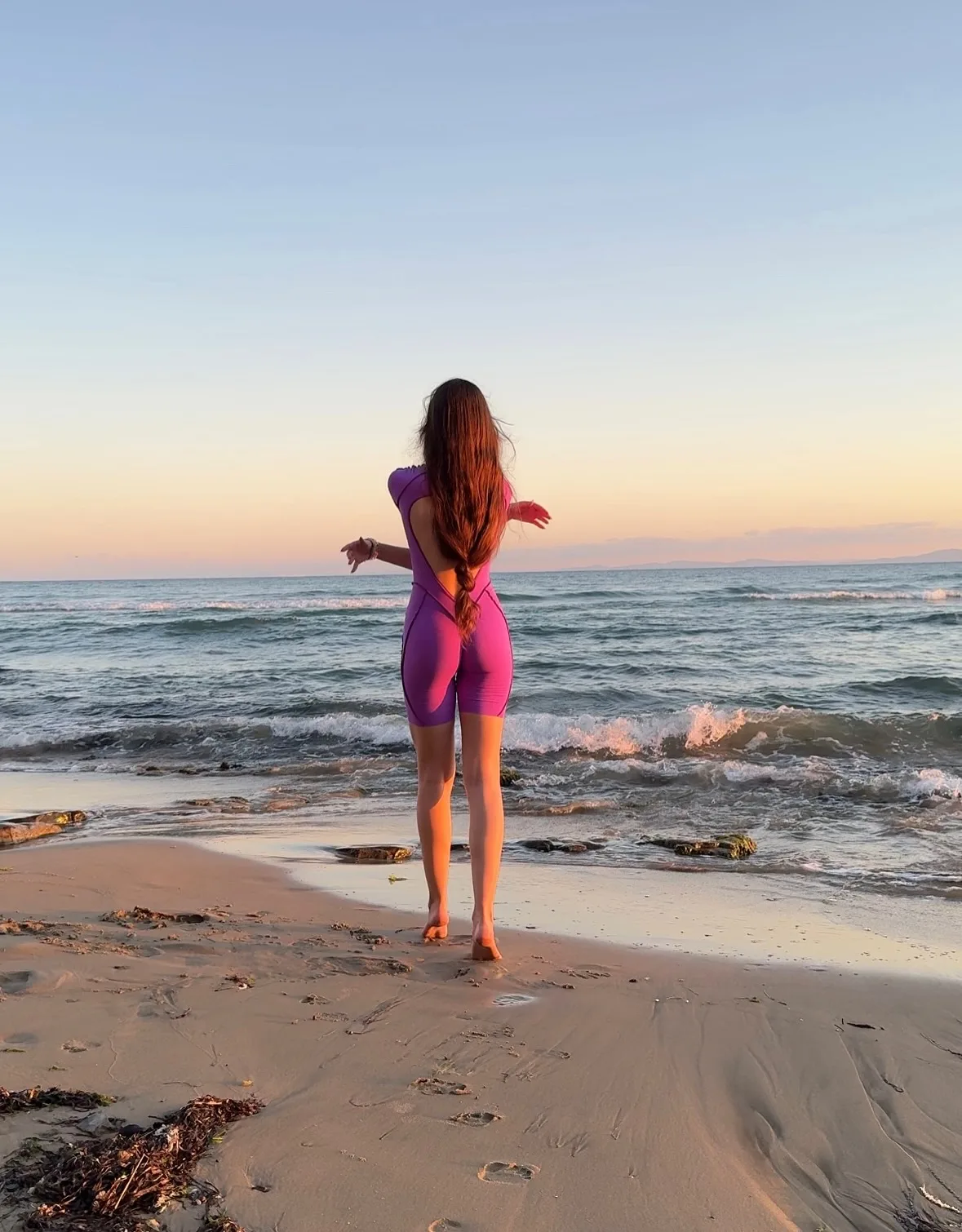 A woman in a purple swimsuit stands on a sandy beach facing the ocean at sunset.
