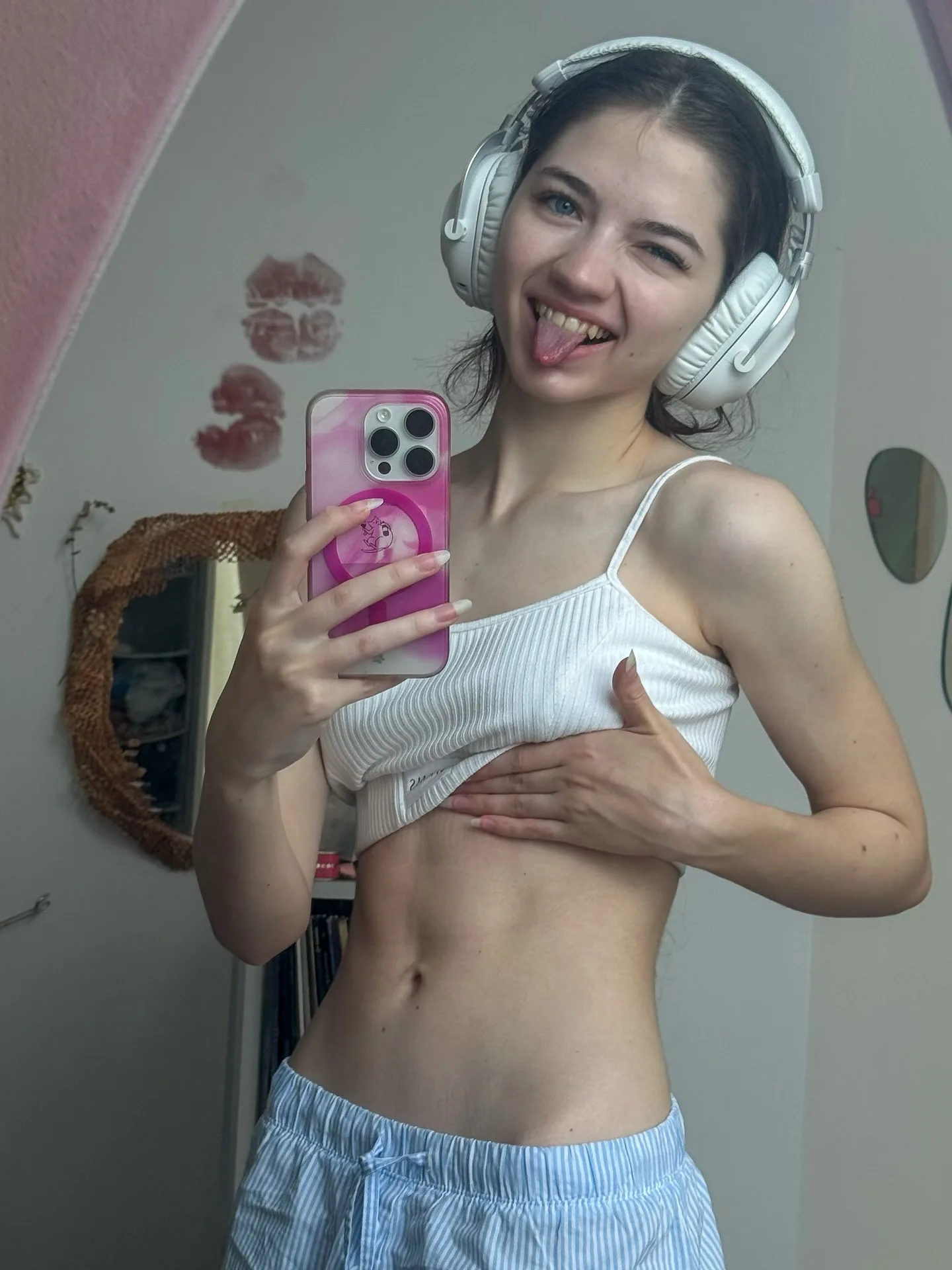 Dana taking a mirror selfie wearing white headphones and a white top, sticking her tongue out while holding a pink phone.
