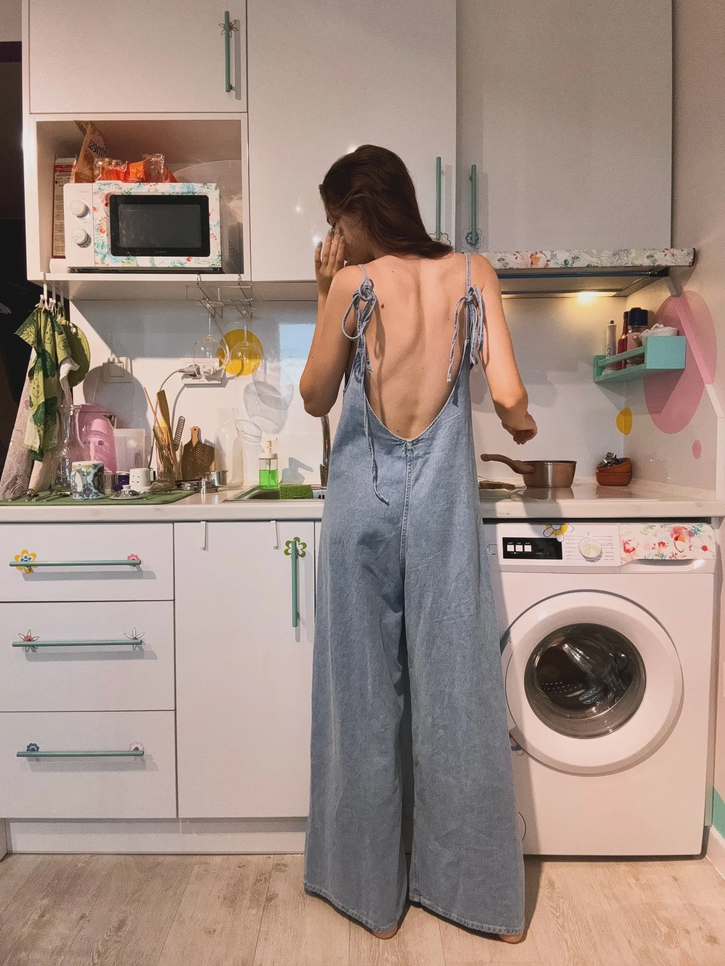 Woman in blue denim overalls with open back standing in a modern kitchen with white cabinets and a washing machine.