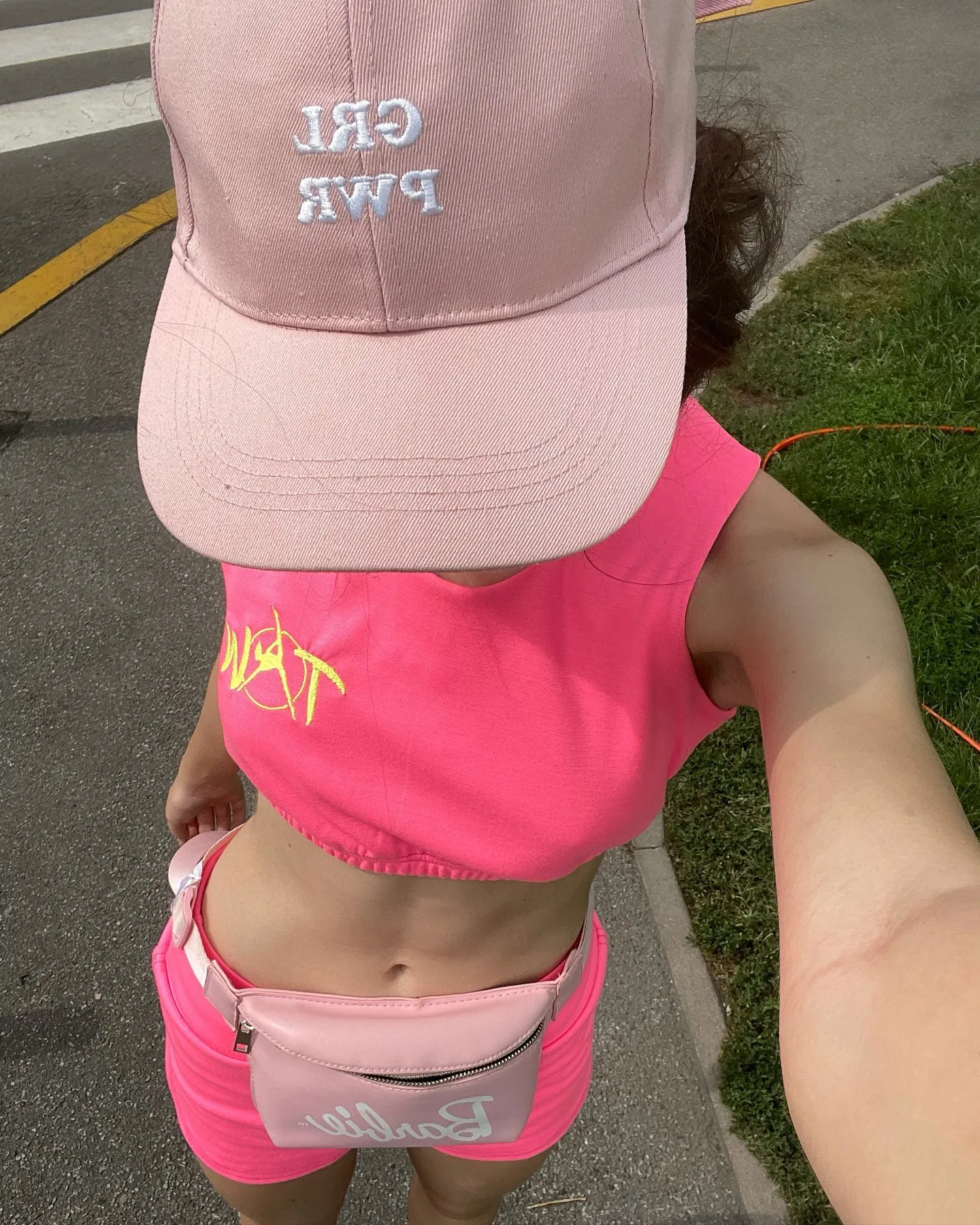 A person wearing a pink cap with "GRL PWR" text, a bright pink crop top, and matching shorts taking a selfie outdoors.