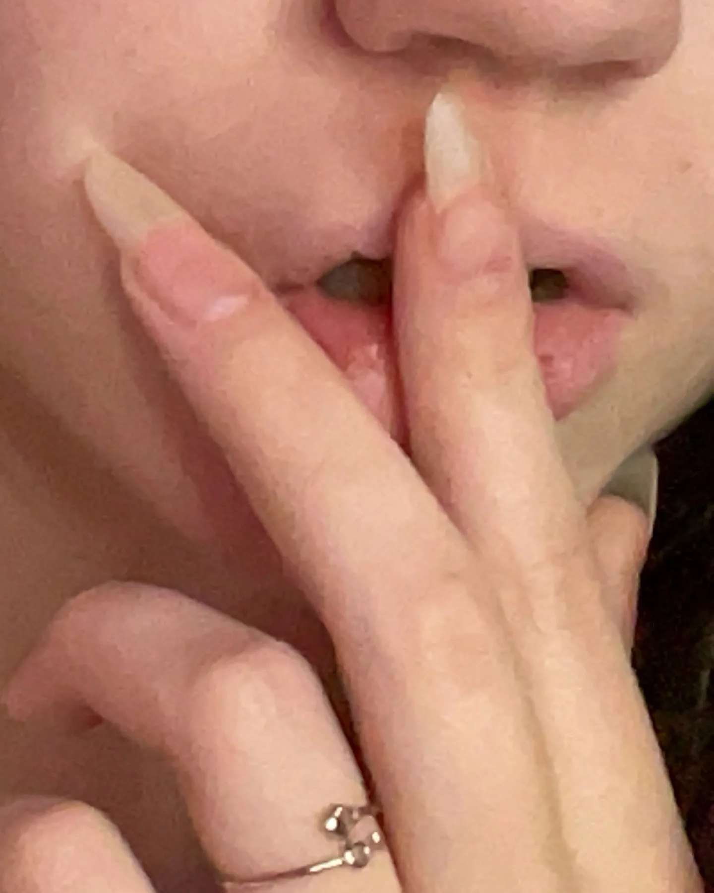 Close-up of a person's mouth and chin with long almond-shaped nails, one finger touching the lip, wearing a delicate ring.