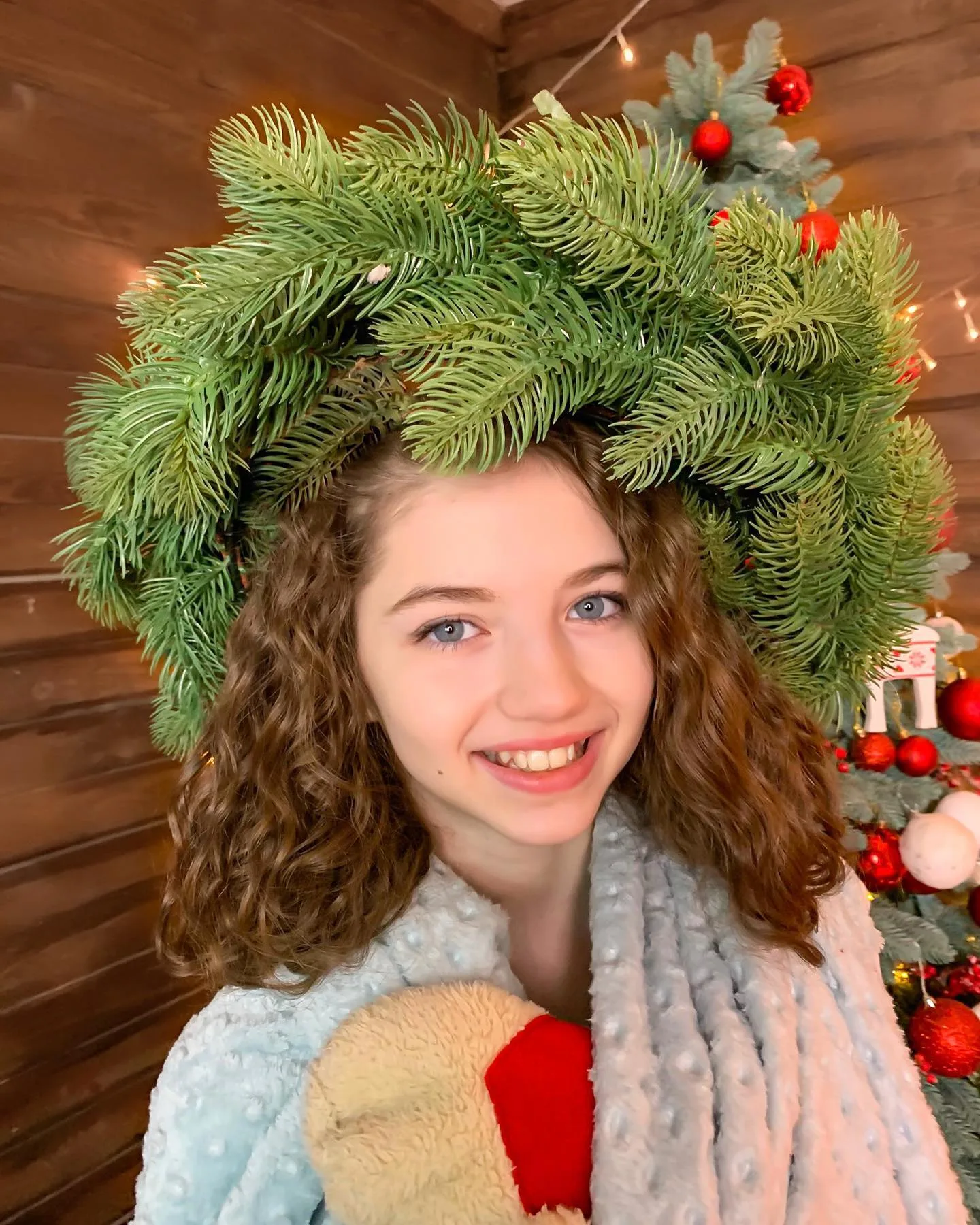 A smiling girl with curly hair wears a large green pine wreath hat and a cozy scarf, holding a gingerbread cookie in a festive wooden room.