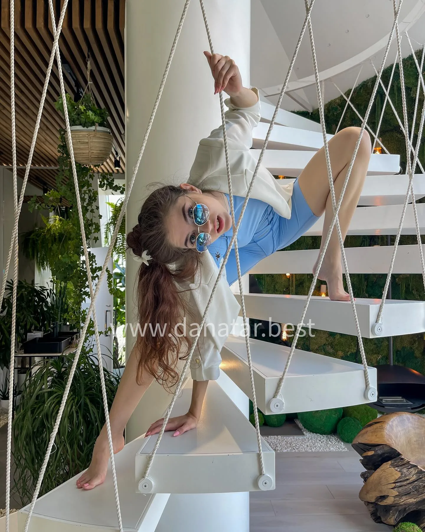 A woman in a white jacket and blue shorts poses playfully on a modern white spiral staircase with rope railings.