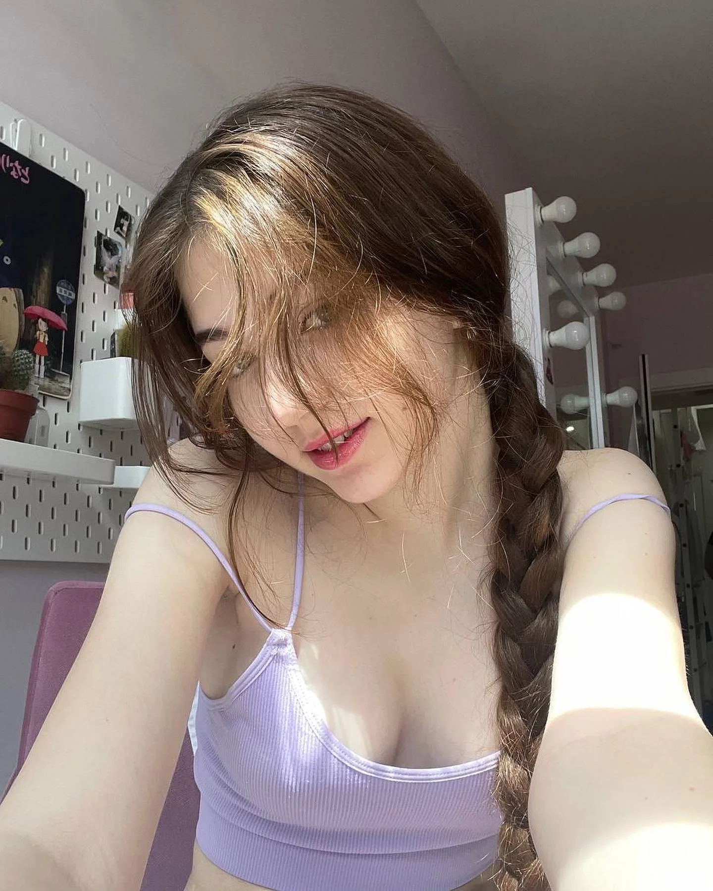 Dana with long brown hair in a side braid wearing a lavender ribbed crop top, posing in a room with a vanity mirror and pegboard wall.