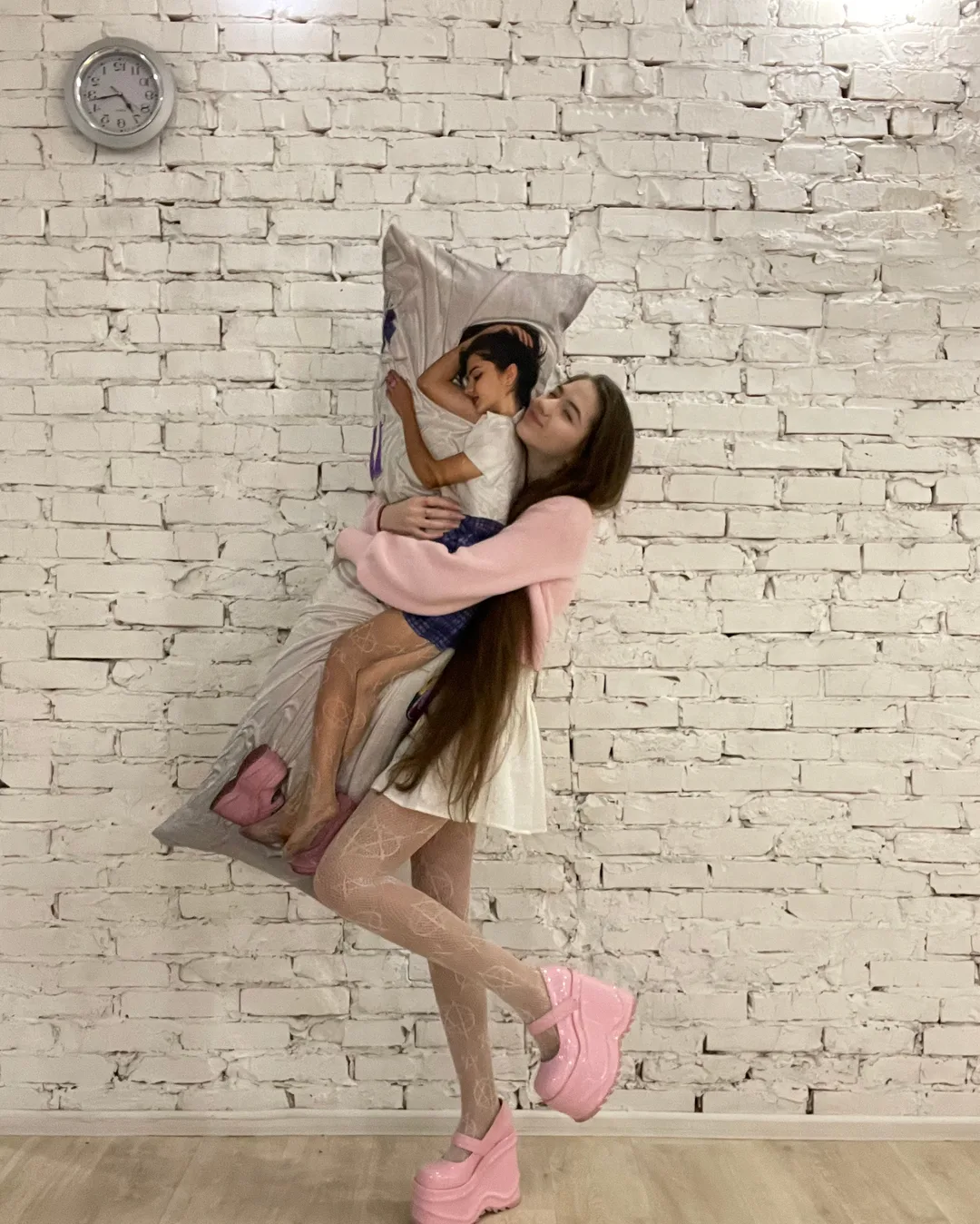 A woman in a pink top and white skirt hugs a large pillow featuring a photo of a child, standing against a white brick wall.