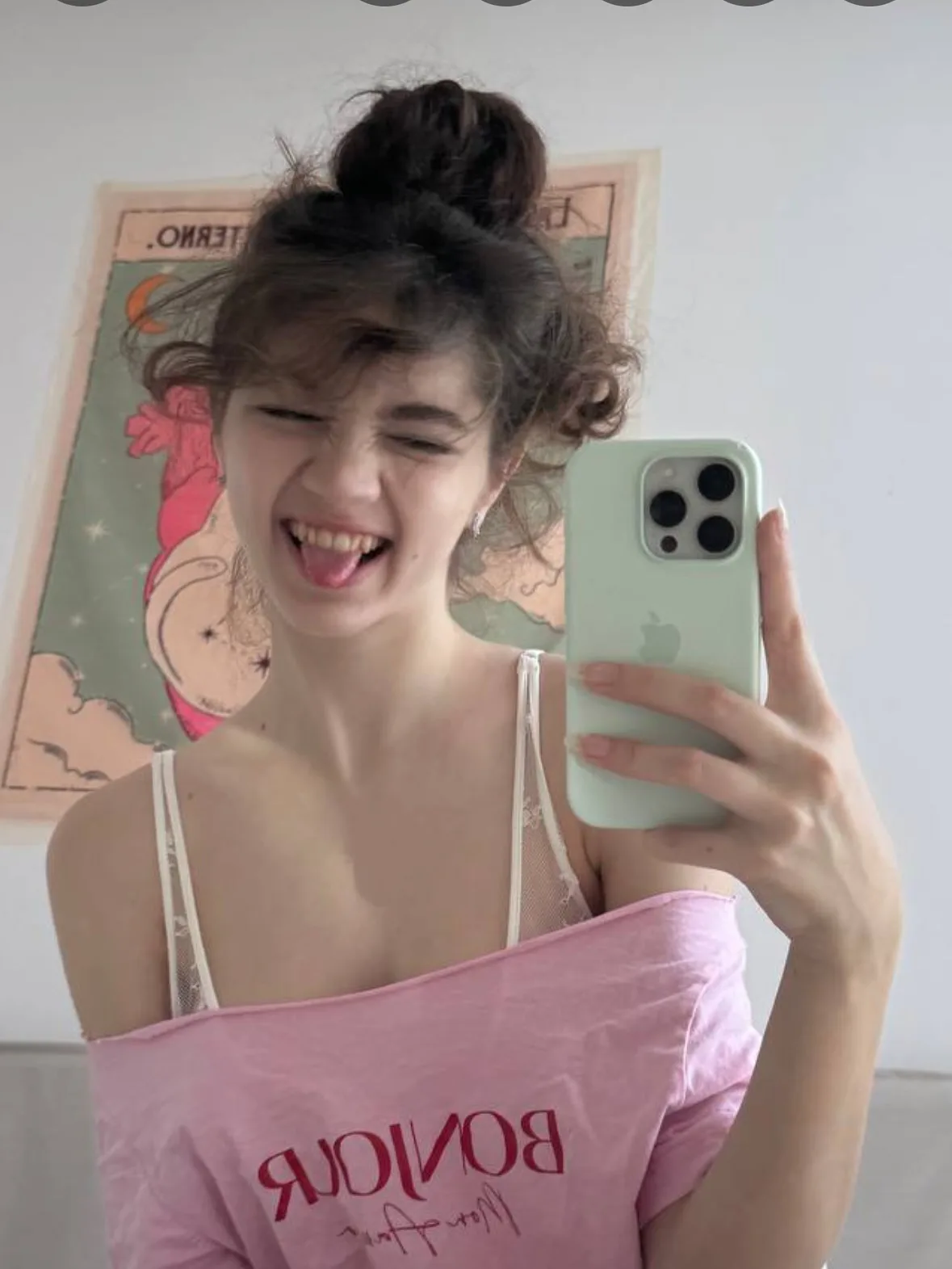 A young woman with curly hair in a bun takes a playful mirror selfie, winking and sticking out her tongue while holding a light green smartphone and wearing a pink top with "BONJOU