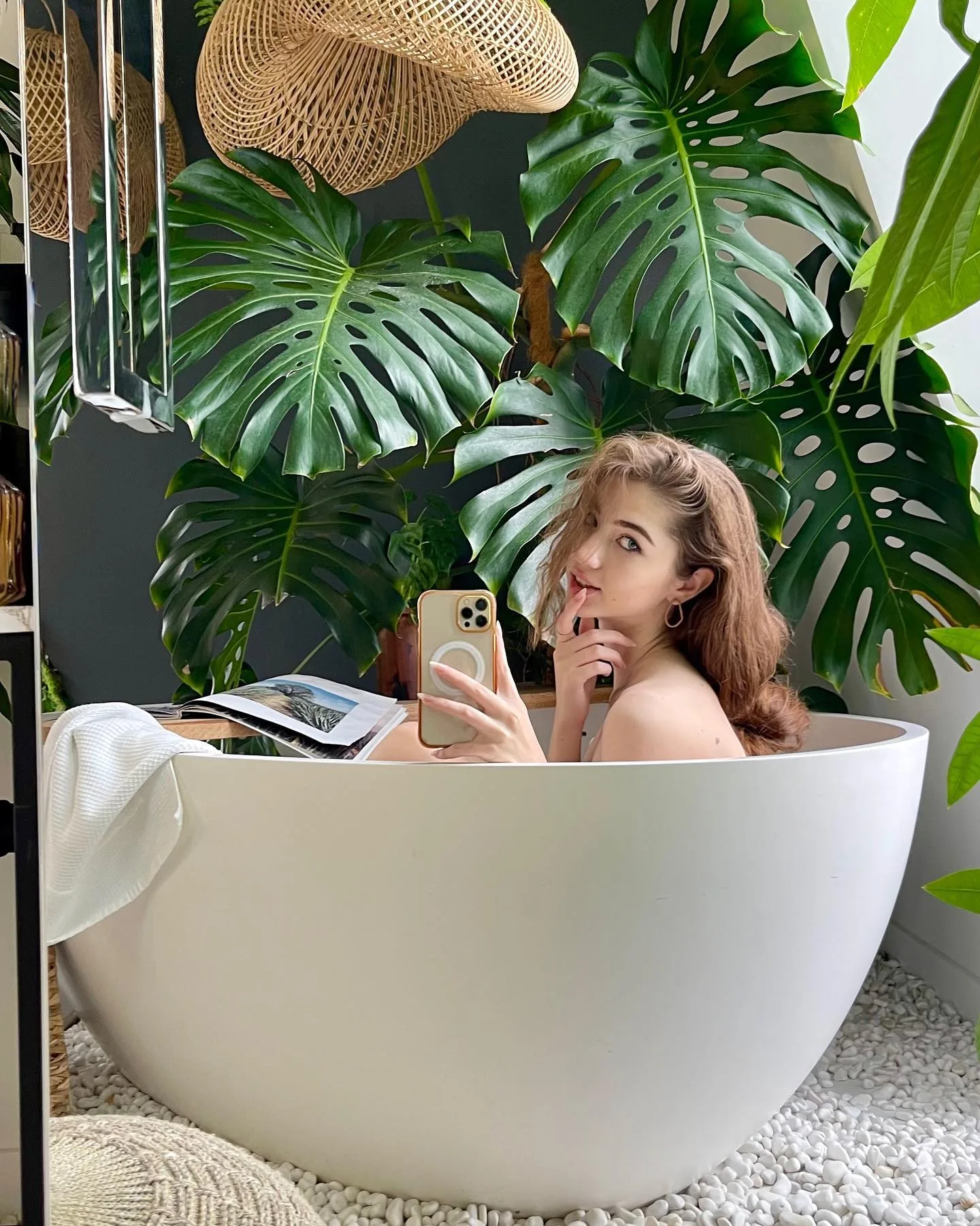 A woman taking a selfie in a white freestanding bathtub surrounded by large monstera plants and a woven hat.