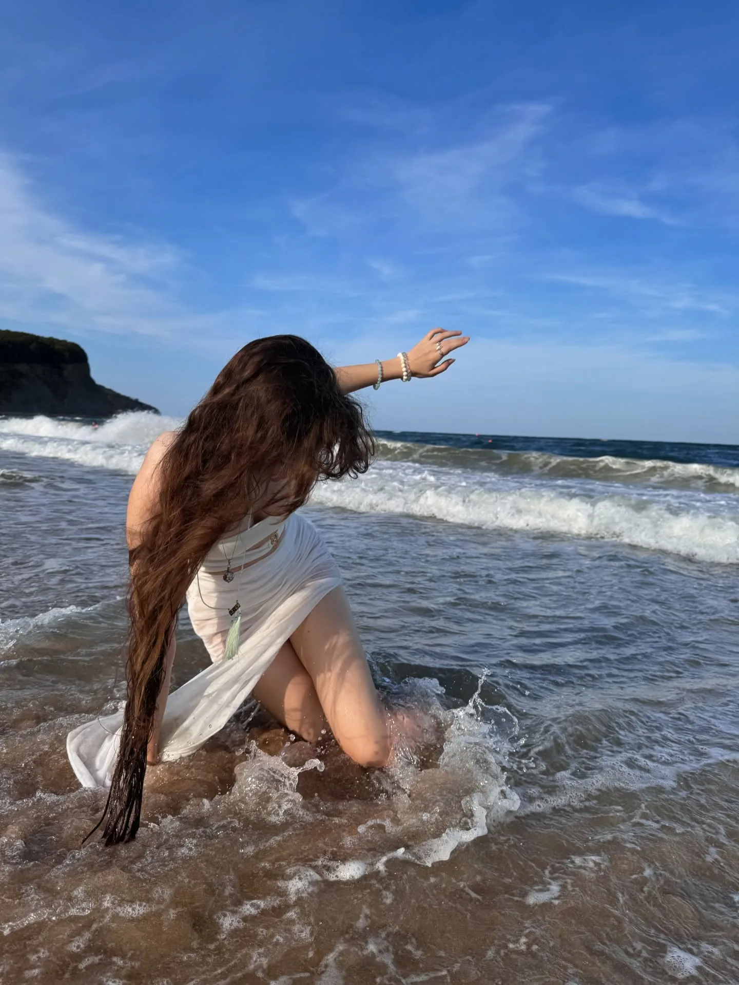 A woman with long dark hair kneels in shallow ocean water, wearing a white dress and reaching out her hand against a blue sky.