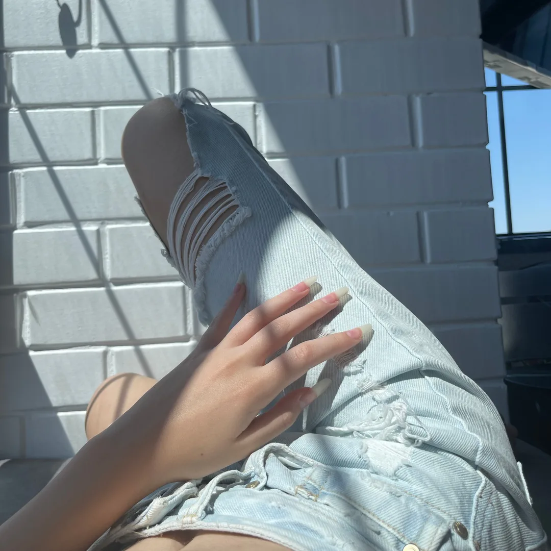 Close-up of a hand resting on ripped denim shorts against a white brick wall.
