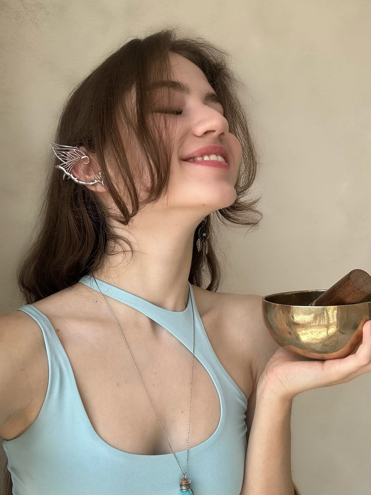 A woman with closed eyes smiles while holding a golden singing bowl, wearing a light blue top and a delicate ear cuff.
