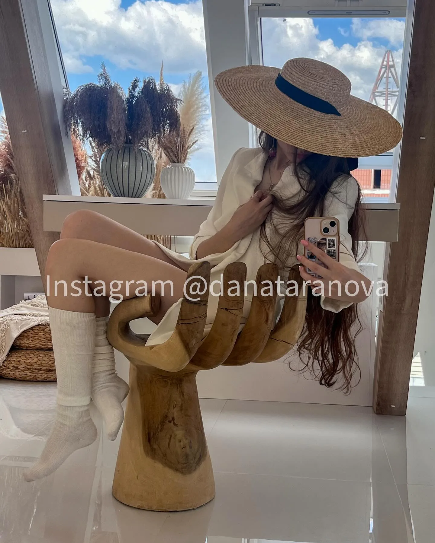Woman in a wide-brimmed hat and white boots taking a mirror selfie while sitting on a wooden hand sculpture.