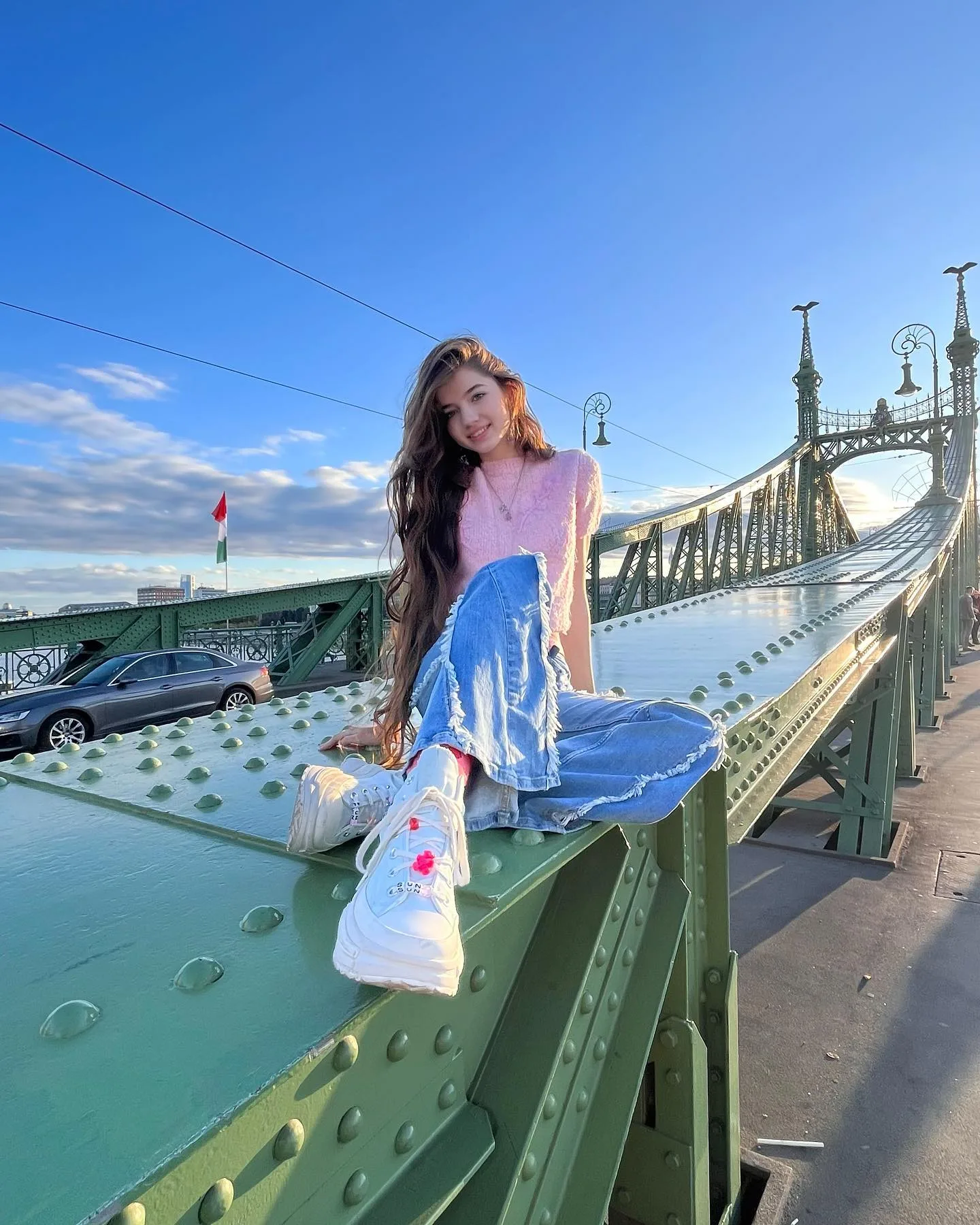 A woman in a pink top and blue skirt sits on the green steel beam of a bridge, holding a white bag against a clear blue sky.