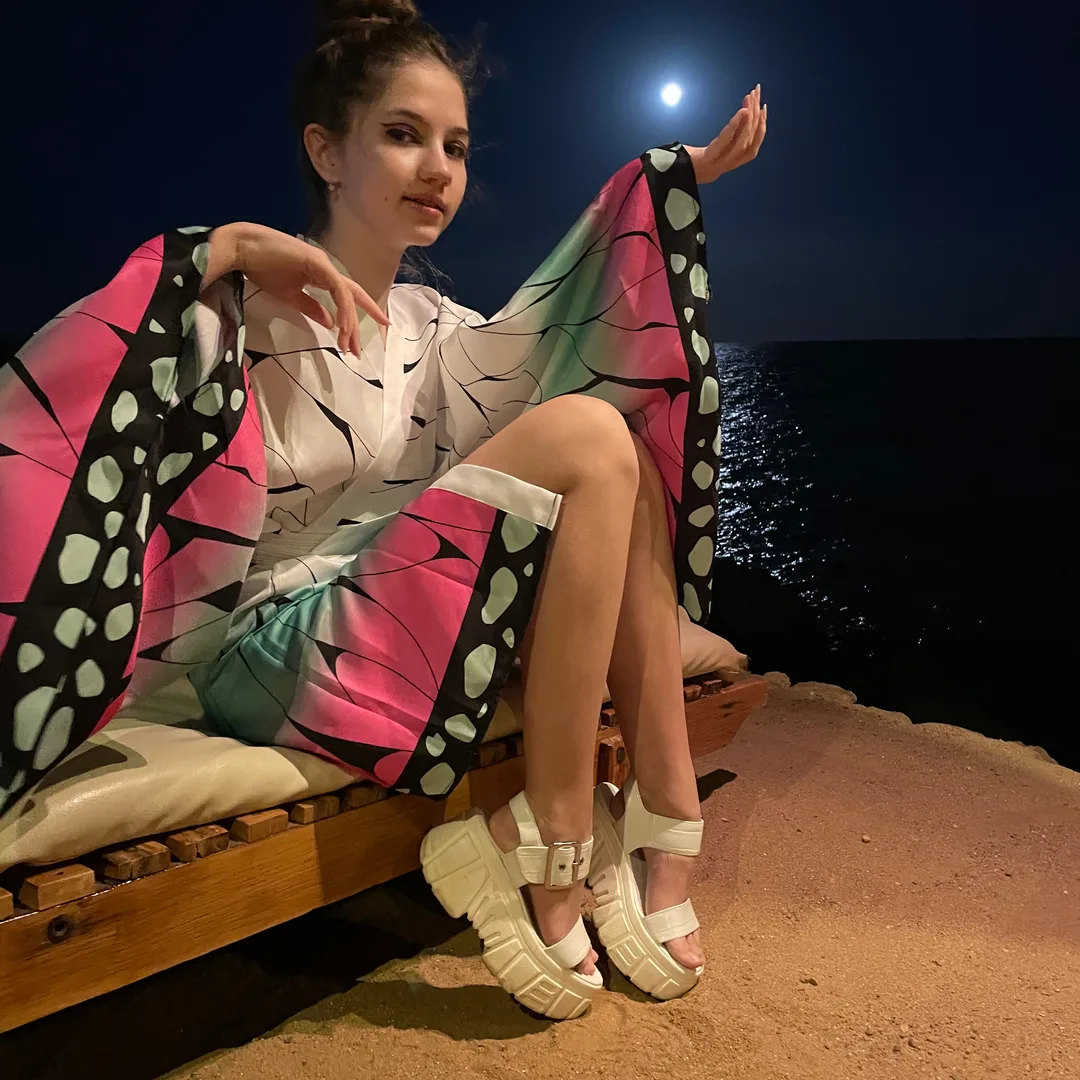 A woman in a colorful butterfly-themed kimono sits on a wooden bench by the sea at night, posing with the moon in the background.